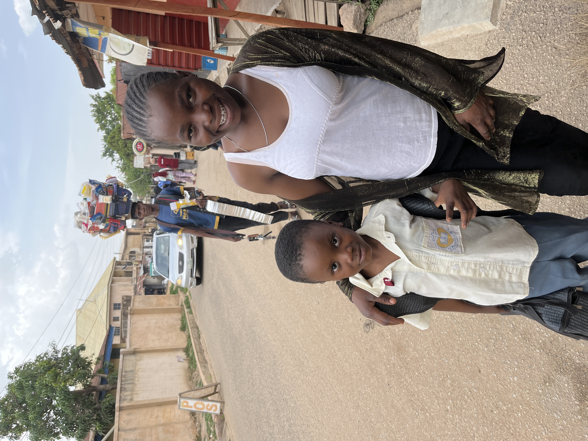 The image captures a woman and a child posing for a photograph on a street in Jos, Nigeria. The woman, with braided hair and a light complexion, is wearing a white tank top with a thin silver necklace, and a shimmering dark shawl. The child, likely a young boy, is wearing a light-colored button-down shirt with a school emblem and dark-colored trousers and has a backpack on. Both are smiling at the camera.
In the background, there is a street scene with several elements. A man is carrying a large, colorful assortment of items on his head. A silver car is passing by, and pedestrians are visible on the sides of the road. Buildings line the street, and a sign labeled "POS" is visible near the bottom of the frame. The environment suggests a typical urban setting in Nigeria.