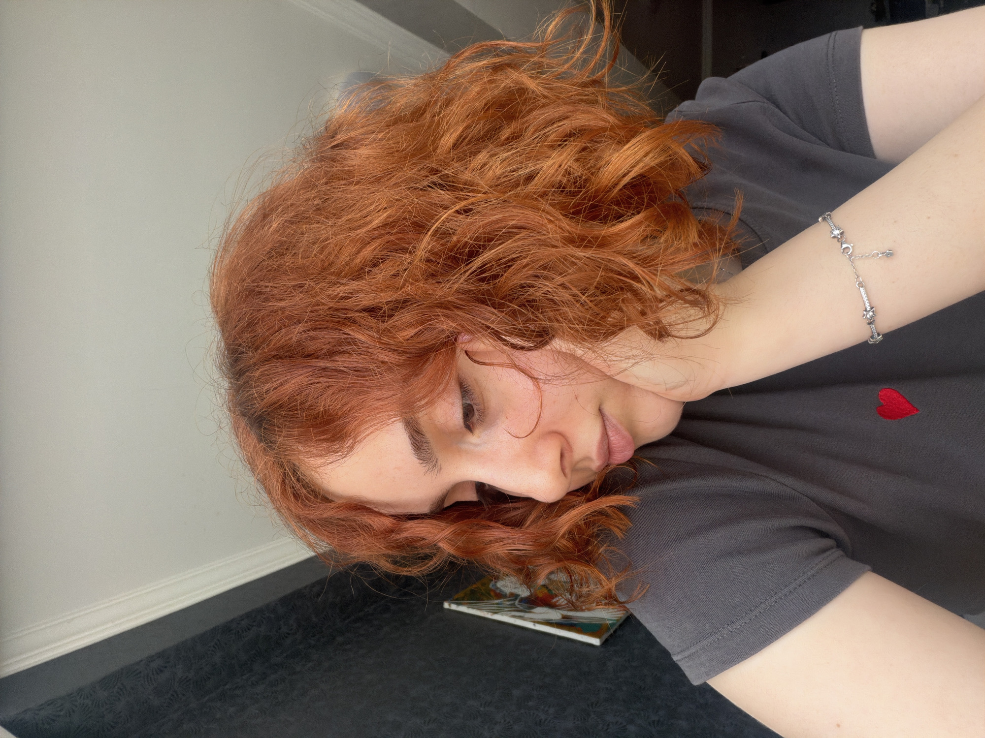A young person with red, curly hair rests their head on a dark surface, seemingly a table or floor, in front of a light gray wall. The individual's face is turned downward and slightly to the side, obscuring the eyes. A dark gray t-shirt with a small red heart emblem is visible on their torso. A silver bracelet adorns their wrist. A small, rectangular object with a colorful design lies underneath their head. The environment consists of a light gray wall and a dark-colored surface underneath the person. The setting does not provide enough information to confirm it is within Tbilisi, Georgia.