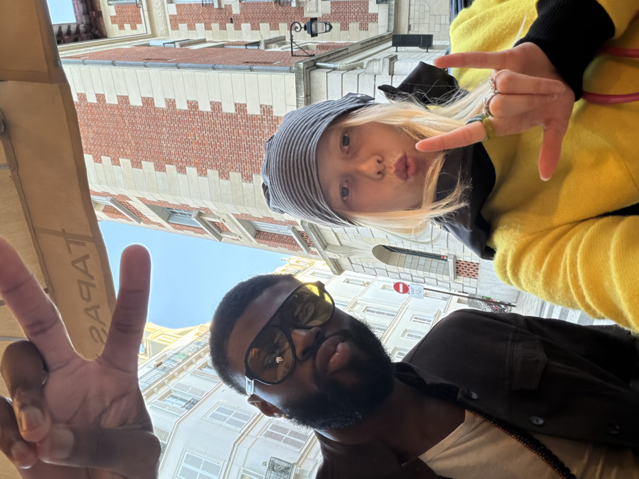 The image is a daytime outdoor selfie featuring two individuals on a street in Paris, France.
In the foreground, a dark-skinned male with a beard and short dark hair is positioned on the left, wearing large, dark-tinted sunglasses and a dark jacket over a white t-shirt. He puckers his lips towards the camera. His right hand, extended into the upper left portion of the frame, forms a peace sign.
To his right, a light-skinned female with blonde hair partially visible under a grey and black striped beanie wears a bright yellow long-sleeved top with a dark collar. She also puckers her lips towards the camera. Her left hand is raised, performing the "rock on" hand gesture, with a green ring on her index finger and other rings on her middle and ring fingers.
The background reveals a typical Parisian urban setting. On the left, a multi-story building with light-colored stone facades, white windows, and black wrought-iron balconies is visible. On the right, a tall building features a distinctive red brick facade with alternating light-colored stone or stucco bands, below which an ornate black street lamp is affixed. A red circular 'Stationnement Interdit' (No Parking) sign is visible on a pole in the mid-ground. Above the subjects, a light-colored awning, partially reflecting the word "TAPAS," is visible against a clear blue sky.