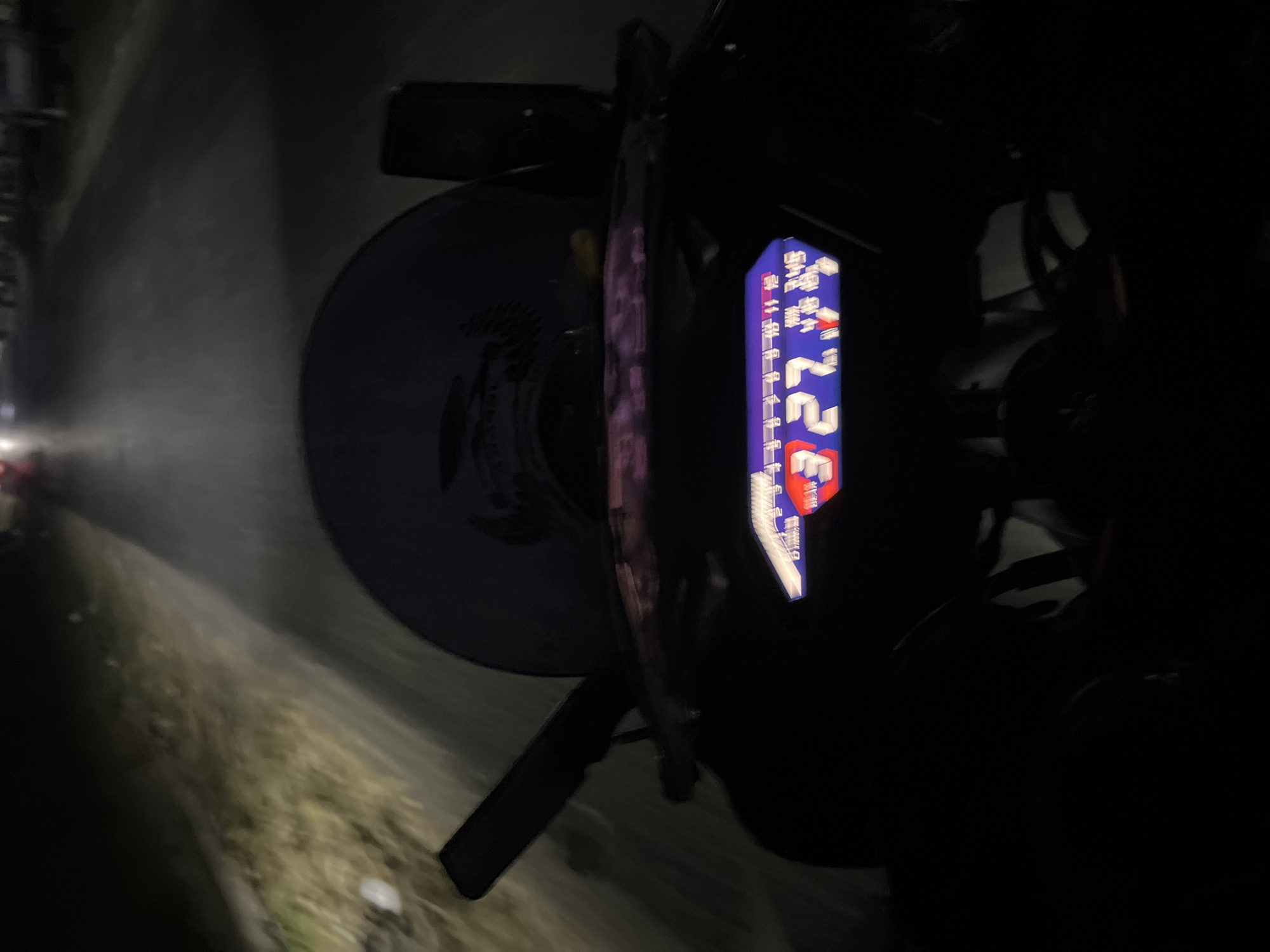 The image captures a first-person perspective of a motorcycle dashboard at night on a road in Karangrandu, Indonesia. The dashboard displays a digital readout, showing the speed as "327" and what appears to be gear indication. The road ahead is visible, illuminated by the motorcycle's headlight and streetlights in the distance, with blurred greenery on the left side. The overall scene suggests movement through a dimly lit urban or semi-urban environment.