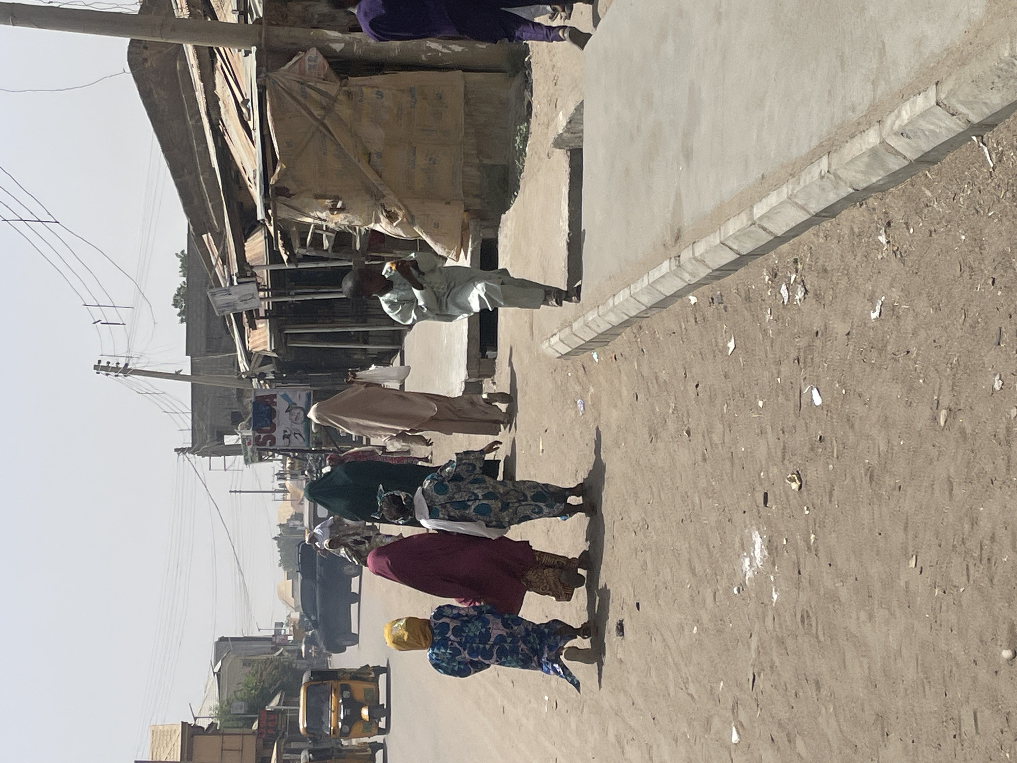 The image depicts a street scene in an urban area. Several people are walking along a dirt road beside a concrete curb. The individuals are wearing a variety of clothing. Buildings and utility poles are visible in the background. Vehicles, including an auto rickshaw, are present on the road.