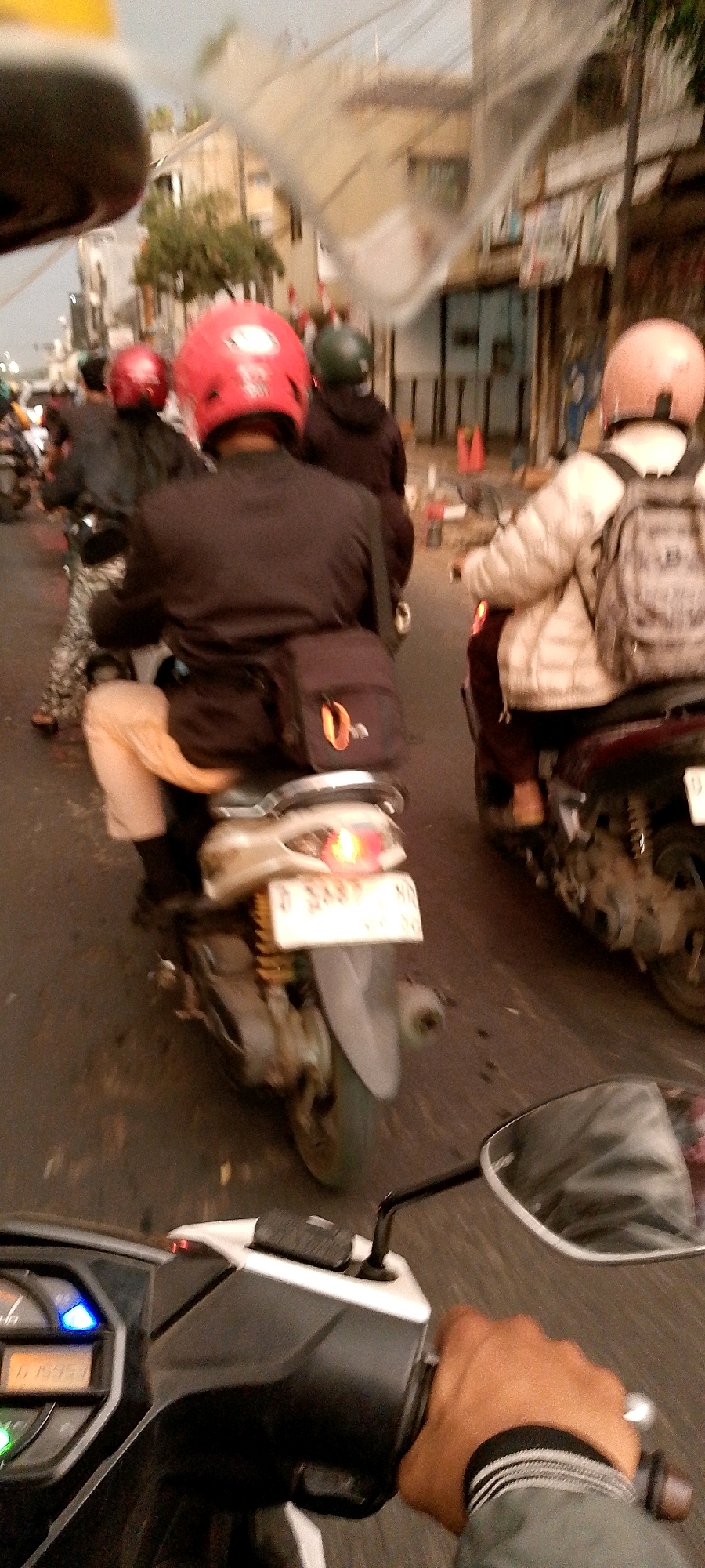 The image depicts a street scene with several motorcyclists in traffic. The perspective is from the rider of a scooter, looking forward and to the right. In the foreground, a hand is visible on the scooter's handlebar, with a partially visible instrument panel showing "6 15953". To the right, the reflection in a scooter's mirror shows a street with buildings and foliage, and another motorcyclist wearing a light-colored jacket and backpack. In the middle ground, a motorcyclist in a red helmet and black jacket is visible, with a brown bag strapped to the back of the scooter. Another motorcyclist in a dark jacket and green helmet is to their left. In the background, the street is lined with buildings and some trees. The overall impression is a busy street scene in an urban environment, possibly Bandung City, Indonesia, given the style of the license plates visible on the scooters.