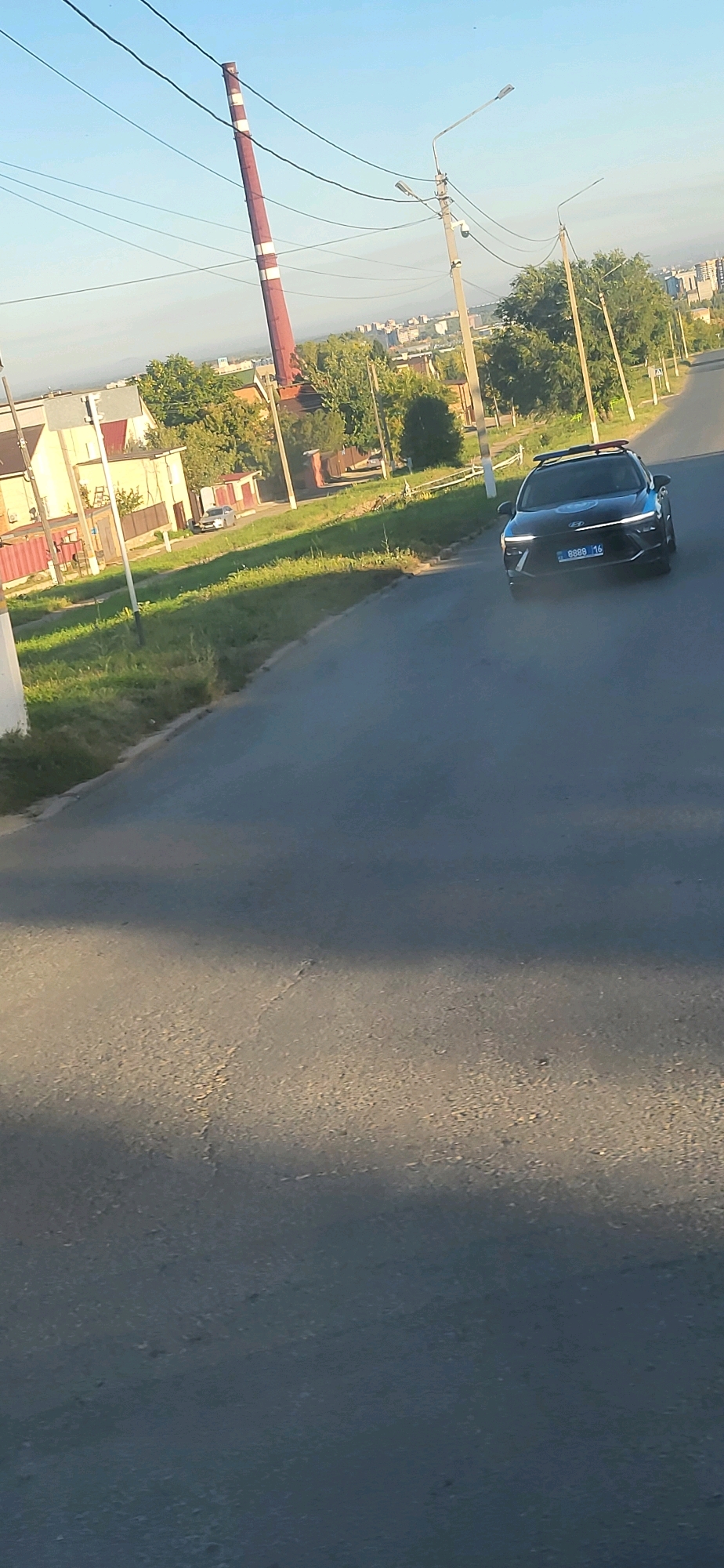 The image depicts a street scene in the city of Oskemen, Kazakhstan. In the foreground, a black Hyundai Accent, possibly a police vehicle due to its light bar, is driving on an asphalt road. The car has the license plate "8888 16". To the left of the road, a patch of grass borders a row of buildings. Further into the background, a tall, red and white striped industrial smokestack stands prominently against a clear blue sky. Trees and other urban structures are visible in the middle and far distance. The lighting suggests it is daytime, likely late afternoon or early evening, given the long shadows cast across the road.