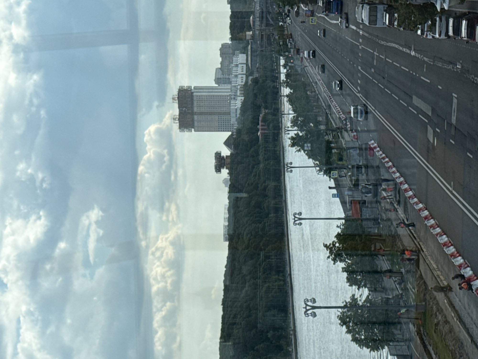 The image presents a wide urban landscape viewed from a high vantage point, likely through a window, evidenced by faint reflections. The upper portion of the frame is dominated by a bright, partly cloudy sky with large, puffy white clouds.
Running horizontally across the mid-ground is a wide body of water, appearing calm and reflective. Along its near bank is a multi-lane asphalt road with visible white lane markings, including solid and dashed lines. Numerous vehicles, including cars and vans, are present on the road, with some moving and others parked. Red and white striped traffic barriers are visible along the road's edge, and a few individuals wearing orange safety vests suggest ongoing construction or maintenance work. Distinctive street lamps with multiple light fixtures are positioned along both the road and the water's edge.
On the opposite side of the water, a dense line of green trees stretches horizontally. Behind these trees, a prominent, tall building with a light-colored facade stands out. It features noticeable structural elements or scaffolding on its upper levels, possibly indicating construction or renovation. Further in the background, other less distinct buildings merge with the distant cityscape. The scene depicts a bustling urban environment during daytime, under fair weather conditions. No legible text is visible.