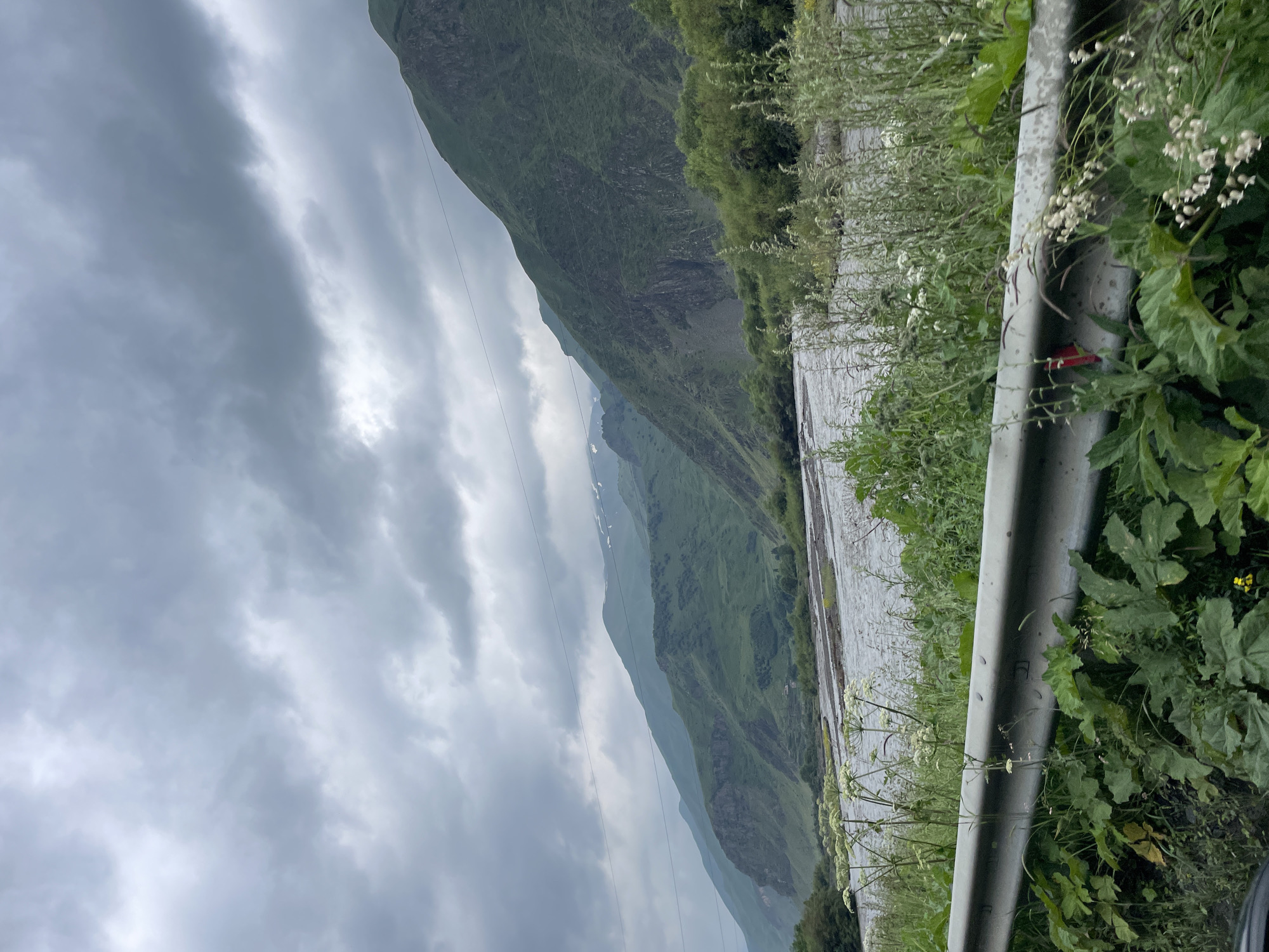 An overcast day in Stepantsminda, Georgia, provides a dramatic backdrop to a mountain landscape. The foreground is dominated by lush green vegetation and a metal guardrail, suggesting the viewpoint is from a road. Beyond this, a wide, shallow river flows through a valley, its surface rippled and gray under the cloudy sky. Steep, verdant mountains rise on either side of the valley, their slopes covered in dense greenery and interspersed with rocky outcrops. In the distance, the peaks of higher mountains are visible, some capped with patches of snow, indicating a high altitude. The sky is a mass of heavy, gray clouds, with hints of lighter patches suggesting the sun is trying to break through. The overall impression is one of a wild, rugged natural beauty, characteristic of the Georgian highlands.