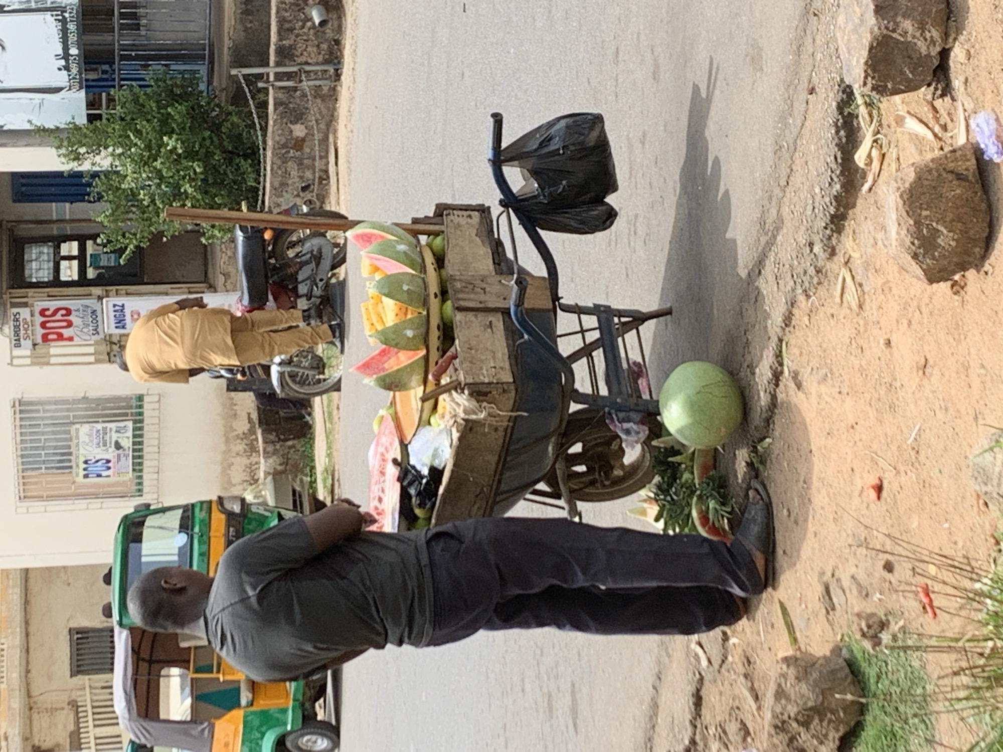 A street scene in Jos, Nigeria. An African male is wearing a gray t-shirt and gray pants and dark footwear stands near a vendor's cart filled with cut watermelon, whole melons, and green fruit, likely avocados. A black plastic bag hangs from the cart. Another African male is standing next to a parked motorcycle, wearing a tan, loose-fitting traditional African robe. A small, open-sided auto rickshaw (tuk-tuk) is parked in the foreground. Buildings are visible in the background. Signs for a Barber Shop and POS Banking Salon are visible.