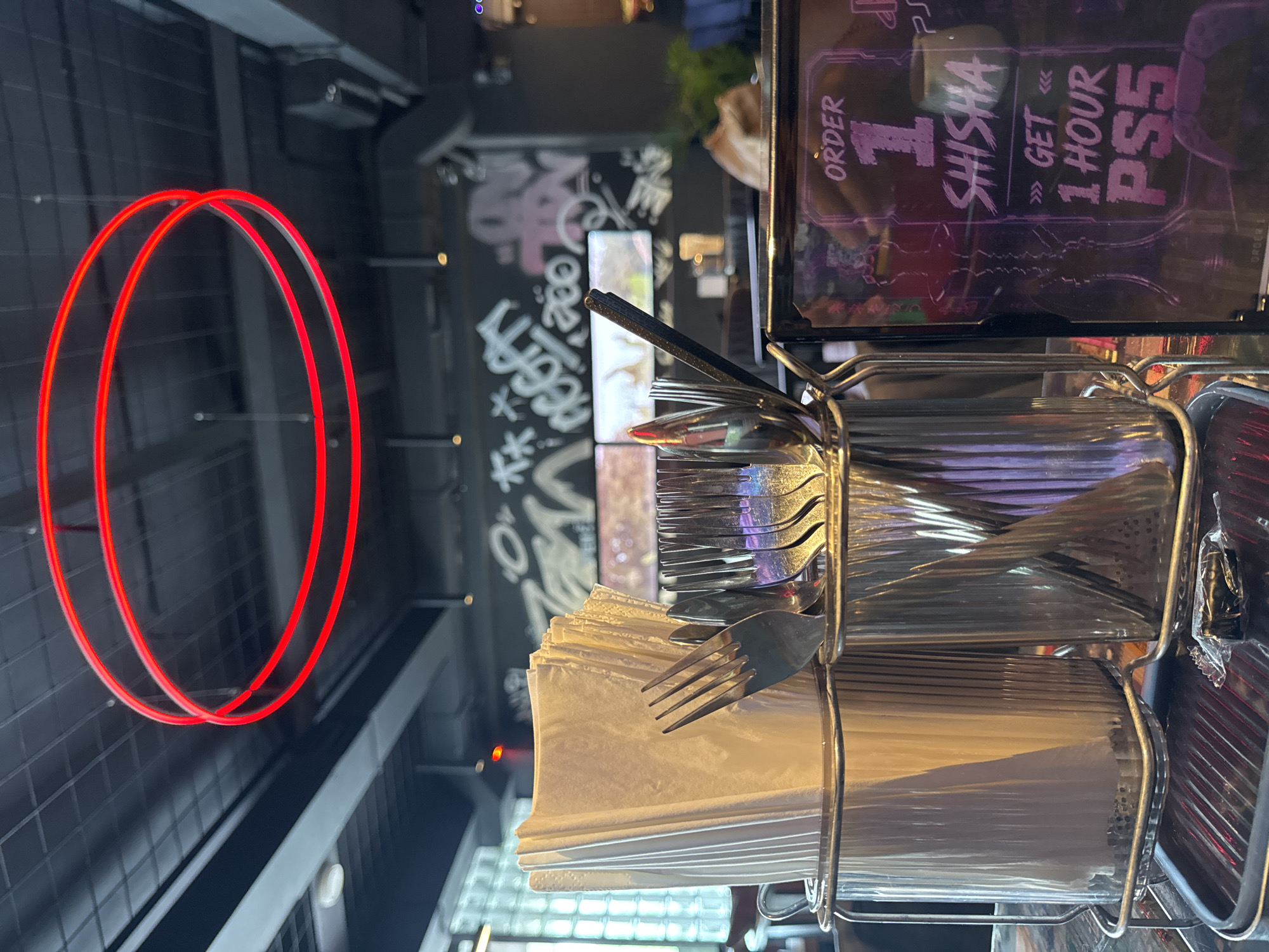 The image depicts a close-up view of a restaurant or lounge setting, likely in Kuta Selatan, Indonesia, with a focus on cutlery and shisha promotion. In the foreground, a metal caddy holds folded white napkins and various utensils, including forks and spoons, all made of stainless steel. To the right, a black framed sign is partially visible, displaying text related to a shisha offer: "ORDER 1 SHISHA GET 1 HOUR PS5". The sign features a silhouette of a shisha pipe and neon-like lettering. The background is blurred, showing a dark interior with graffiti on the wall and what appear to be screens or displays on the wall. Above, red neon lights form concentric circles, suggesting a modern and possibly trendy atmosphere. The lighting is dim, with artificial lights highlighting the foreground objects and the neon accents. No people are clearly visible in the frame.