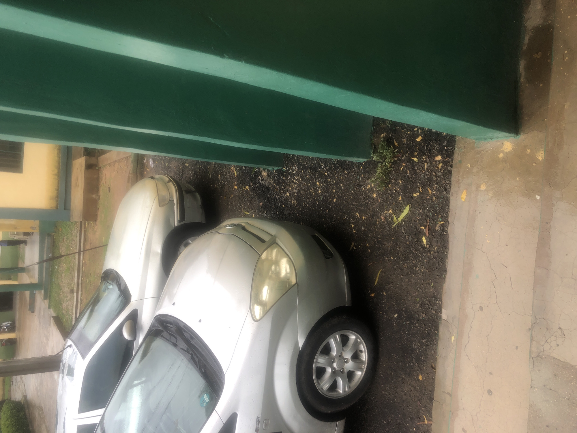The image depicts two silver cars parked next to each other on a dark, gravelly surface, possibly a street or parking lot in Jalingo, Nigeria. The car in the foreground is a silver hatchback, while the car behind it appears to be a silver sedan. A tall, bright green structure, possibly a building's facade or a partition, dominates the right side of the frame, casting shadows on the ground. The environment suggests an outdoor setting, with some fallen leaves scattered on the ground. The lighting indicates it might be daytime, possibly overcast or rainy, as the surfaces appear damp and the light is diffused. There are no people visible in the immediate vicinity of the cars or the green structure. The scene is devoid of any discernable activity.