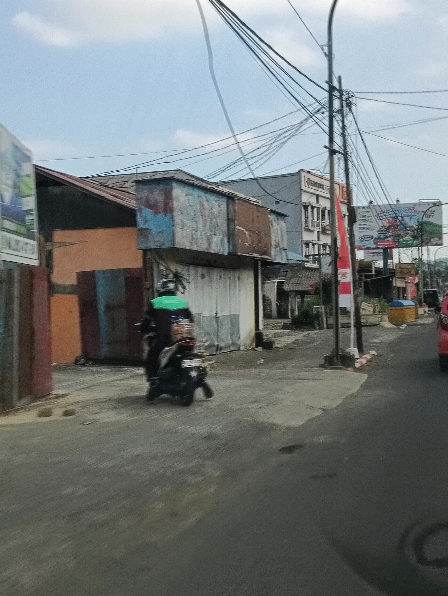The image depicts a street scene in Tomohon, Indonesia. In the foreground, a person wearing a green vest rides a motorcycle, which is blurred due to motion. The motorcycle has a brown box attached to the back. The street is paved with asphalt. To the left, there are buildings with corrugated metal roofs and faded paint. One building has a red and brown facade. Further back, a multi-story building with white and grey sections is visible, with a prominent pole supporting numerous electrical wires overhead. A large billboard displaying text and images is mounted on the right side of the street, near the multi-story building. A red and white flag is also attached to a pole. Several other structures, including what appear to be small shops or stalls, line the street. The sky is light blue with some clouds.