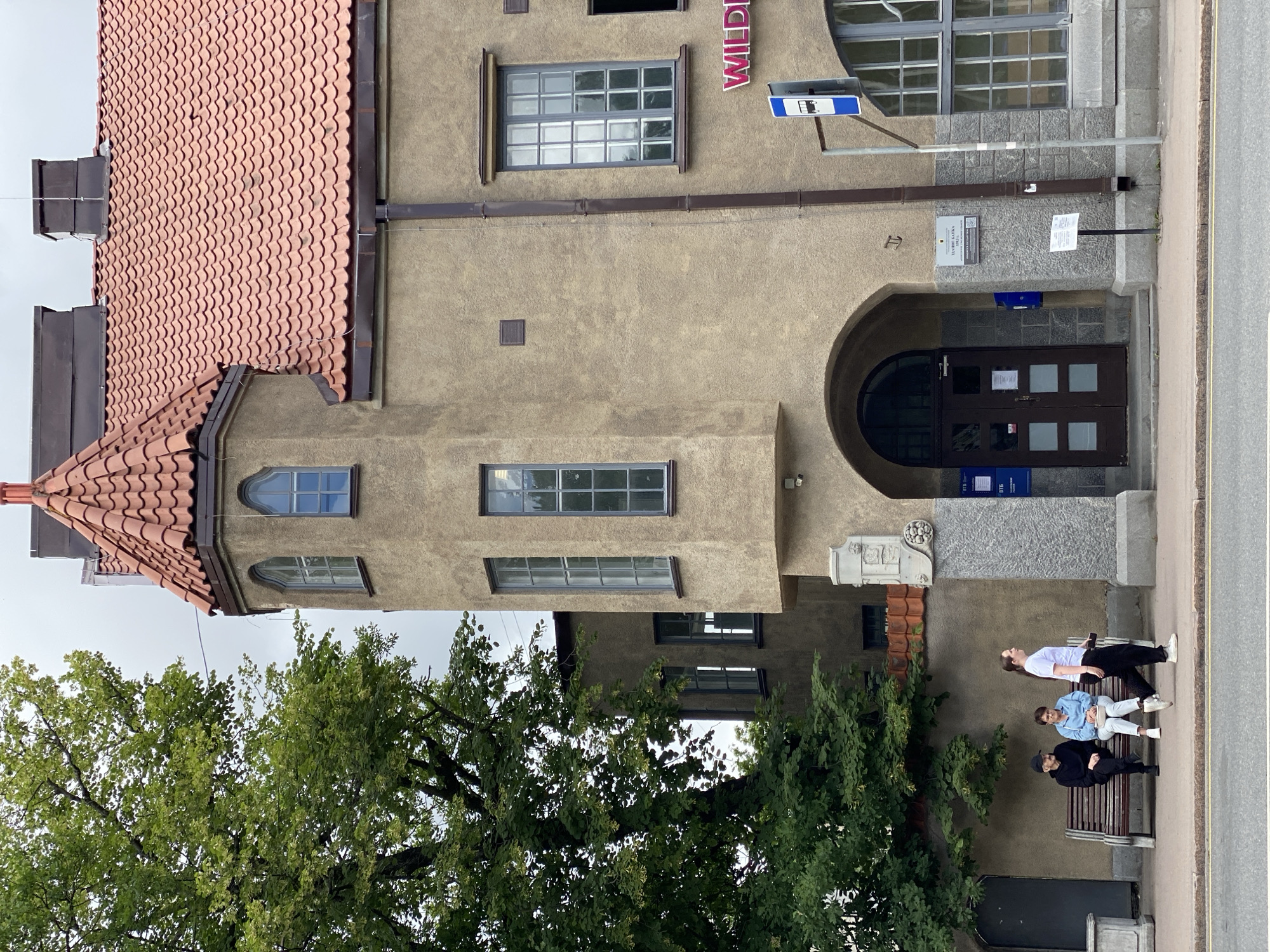 The image shows a street view in Sortavala, Russia, focusing on the exterior of a tan-colored building with a red tile roof and a prominent turret. In front of the building, there is a sidewalk with a bench where two individuals are seated, and another person is walking past them. The building features arched doorways, multiple windows, and a sign that appears to read "WILD." A bus stop sign is visible on the right side of the image. Lush green trees are present on the left, partially obscuring the building. The sky is overcast.
