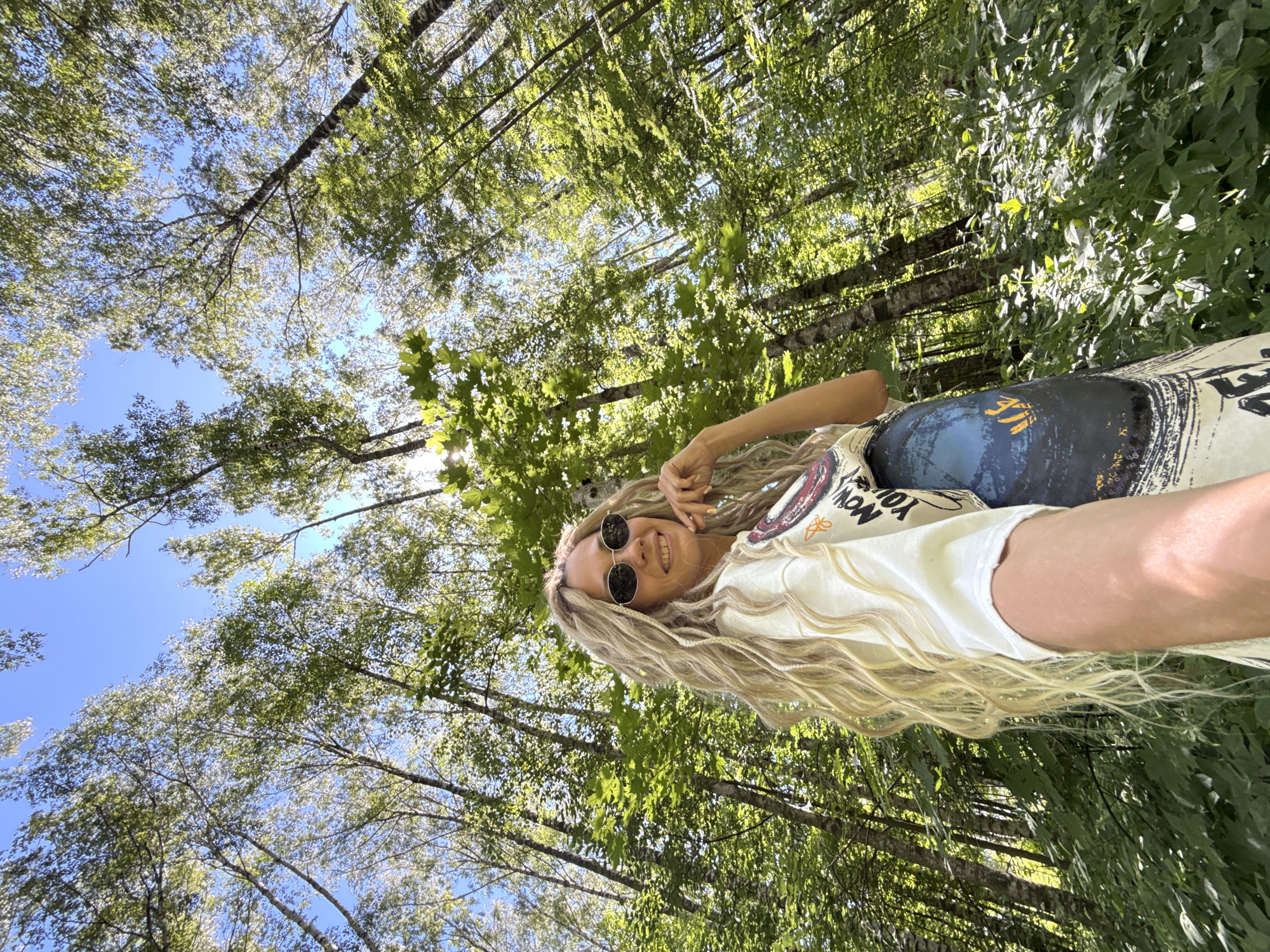 An outdoor, low-angle selfie captures a female subject with long, blonde, textured hair and dark, round sunglasses. She is smiling and positioning her left hand on her chin. The subject wears a light-colored t-shirt featuring a graphic design partially visible as "NOW YOU" and a darker garment with a blue and orange pattern. The background consists of a dense forest with tall trees, predominantly deciduous, displaying vibrant green foliage that extends towards a clear blue sky. Sunlight filters through the canopy, creating bright areas on the leaves and illuminating the scene. The ground is covered with green undergrowth. The image depicts a sunny day in a wooded area within the city of Rusanovo, Russia.
