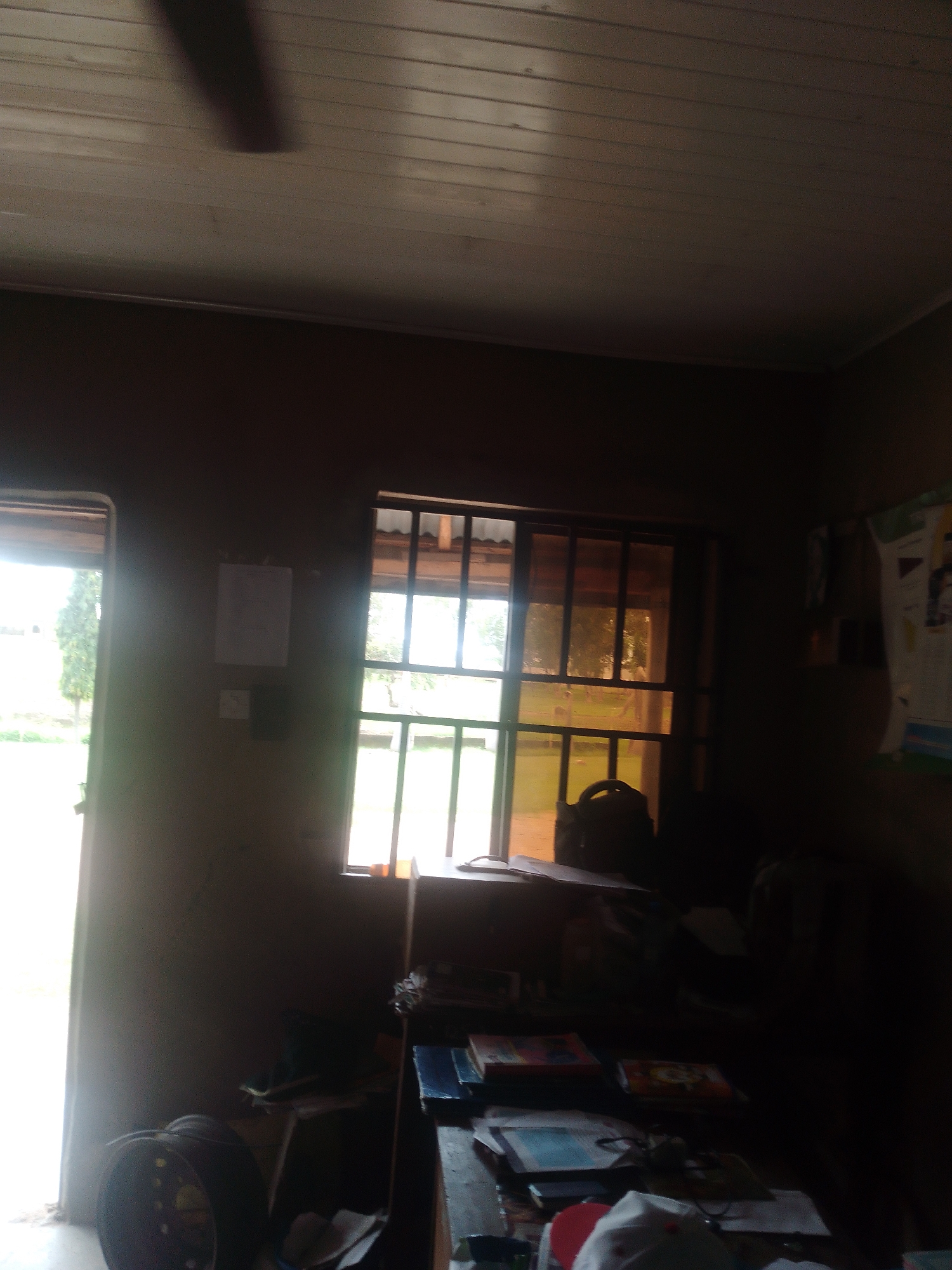The image depicts a dimly lit room with two windows. One window, on the left, is open, revealing an outdoor scene with trees and what appears to be a grassy area. Sunlight streams in from this window. The other window, to the right, is partially obscured by blinds or bars and is illuminated by an amber-toned light, suggesting it might be late afternoon or evening, or that the window has tinted glass.
Inside the room, there is a ceiling fan, partially visible at the top of the image, with one of its blades casting a shadow. On the wall next to the left window, there are two pieces of paper or documents attached. To the right of the right window, a large poster or notice board with some text and images is hung on the wall.
In the foreground, a desk or table is visible, cluttered with books and papers. Some of the books appear to be stacked. A dark-colored bag or backpack is placed on a surface near the right window. On the far left of the image, near the open doorway, a large, dark, circular object, possibly a tire, is leaning against the wall.
The overall impression of the room is one of a functional space, perhaps an office or study, with an academic or informational display on the wall. The lighting suggests it is either daytime with an overcast sky, or the room itself is not well-lit, with the main light source coming from the open window. There are no people visible in the image. The location context of Jauro Yinu, Nigeria, provides a geographical anchor for the scene.