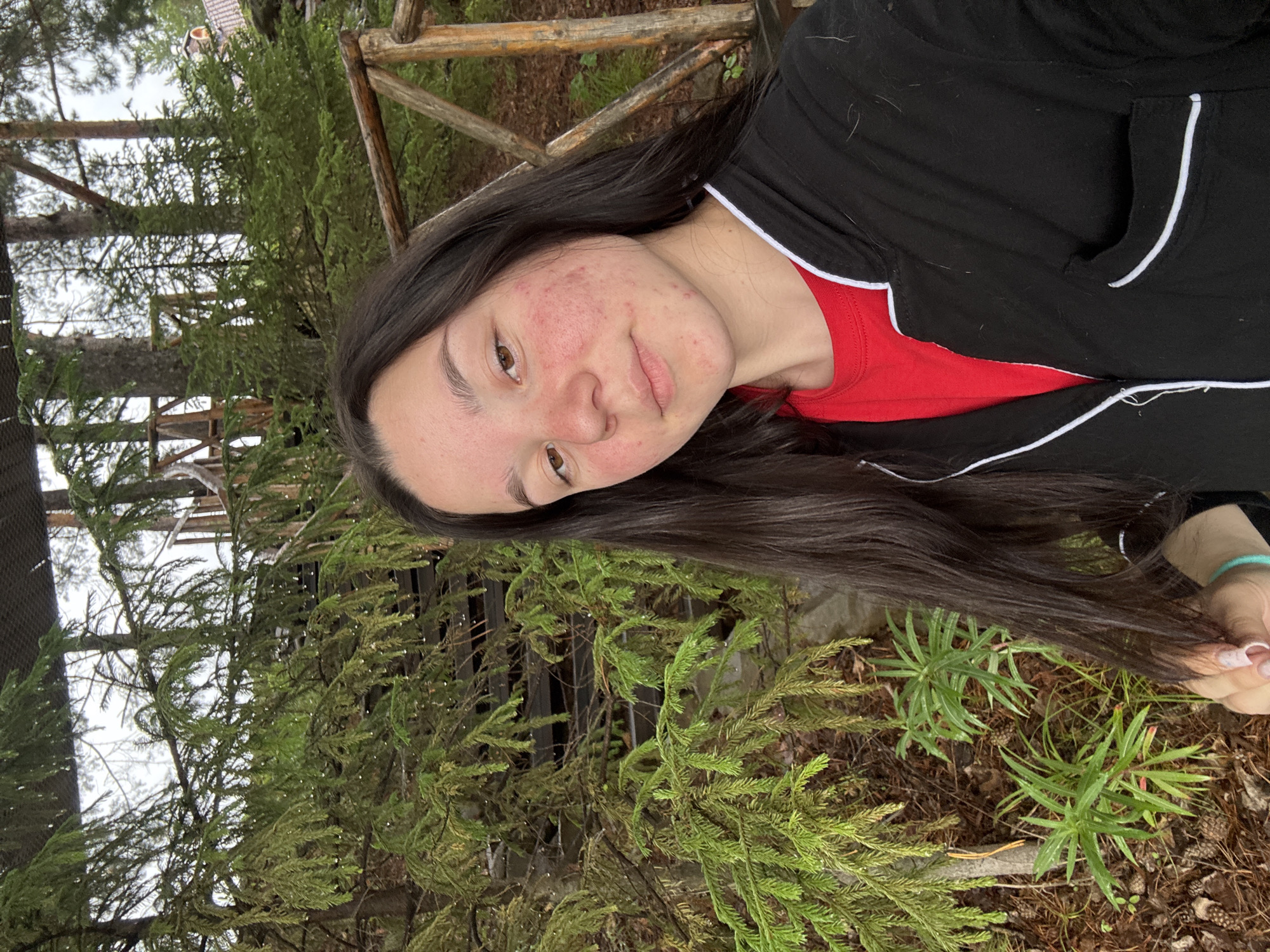 The image shows a young woman with long, dark hair and fair skin, turned towards the camera with her head tilted. She is wearing a red t-shirt and a black jacket with white trim. Her face has some redness and blemishes, and she has brown eyes and a slight smile. She is outdoors, with a background of pine trees and a rustic wooden structure that appears to be a fence or railing. The ground is covered with pine needles and small green plants. The lighting suggests it might be daytime, possibly overcast or in a shaded area due to the soft light. The overall impression is a casual, outdoor portrait taken in a natural, wooded setting. The location is identified as Pozdyshki, Russia. There is no visible text in the image.
