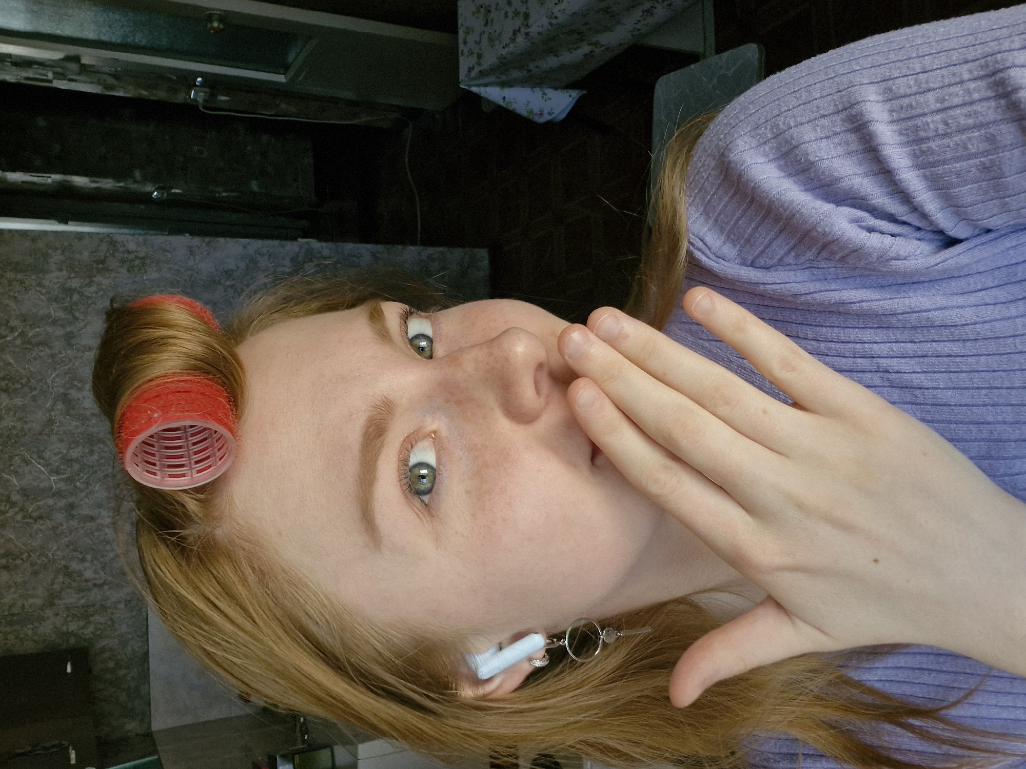 A fair-skinned woman with light brown hair, freckles, and green eyes is depicted. One red hair roller is visible on the left side of her head. She is wearing a light purple, horizontally ribbed shirt or sweater. Her hand is held up to her face, partially covering her mouth. She wears a unique earring that appears to have a white item inserted in it, likely a wireless earbud, connected to a metal loop. The background shows a kitchen or bathroom counter with dark cabinets and a patterned wall, indicative of a domestic interior. Given the prompt's request, this scene does not appear to be in Yekaterinburg, Russia based on the limited visible context. There are no obvious indicators such as unique architecture or cultural symbols that would suggest that location.
