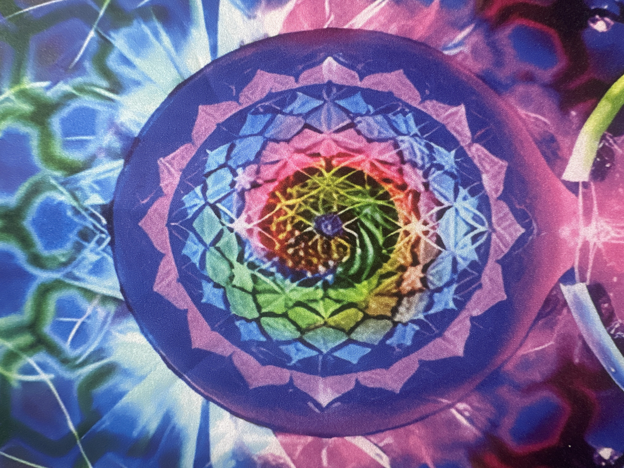 The image shows a close-up of a colorful, intricate circular pattern, possibly a tapestry or a printed fabric. The pattern features a mandala-like design with multiple concentric layers of geometric shapes and vibrant colors, including blues, purples, pinks, greens, and yellows. The central area displays a swirling vortex of rainbow hues, surrounded by diamond and petal-like shapes that transition in color. The outer portions of the image show abstract, energetic patterns in shades of blue, green, and pink, with some white light effects radiating outwards. The texture of the fabric is visible, indicating it is a soft material. There are no people, specific landmarks, or any indication of its location within Letterkenny, Ireland.