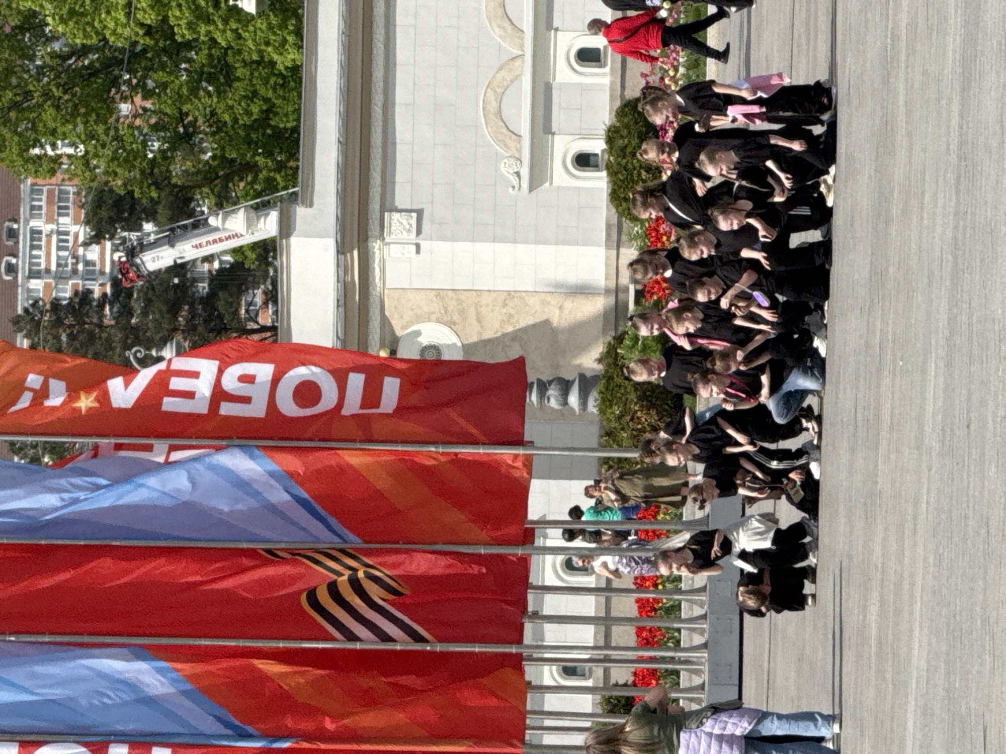 A side-angle outdoor shot shows a group of children wearing black, standing on a wooden deck in front of red and blue flags with black-and-orange ribbons. The flags read "Победа" (Victory) in Russian. Behind them, there is a building with a fire truck lift. The setting is likely Moscow, Russia, based on the language on the flags. The children appear to be attending an event.