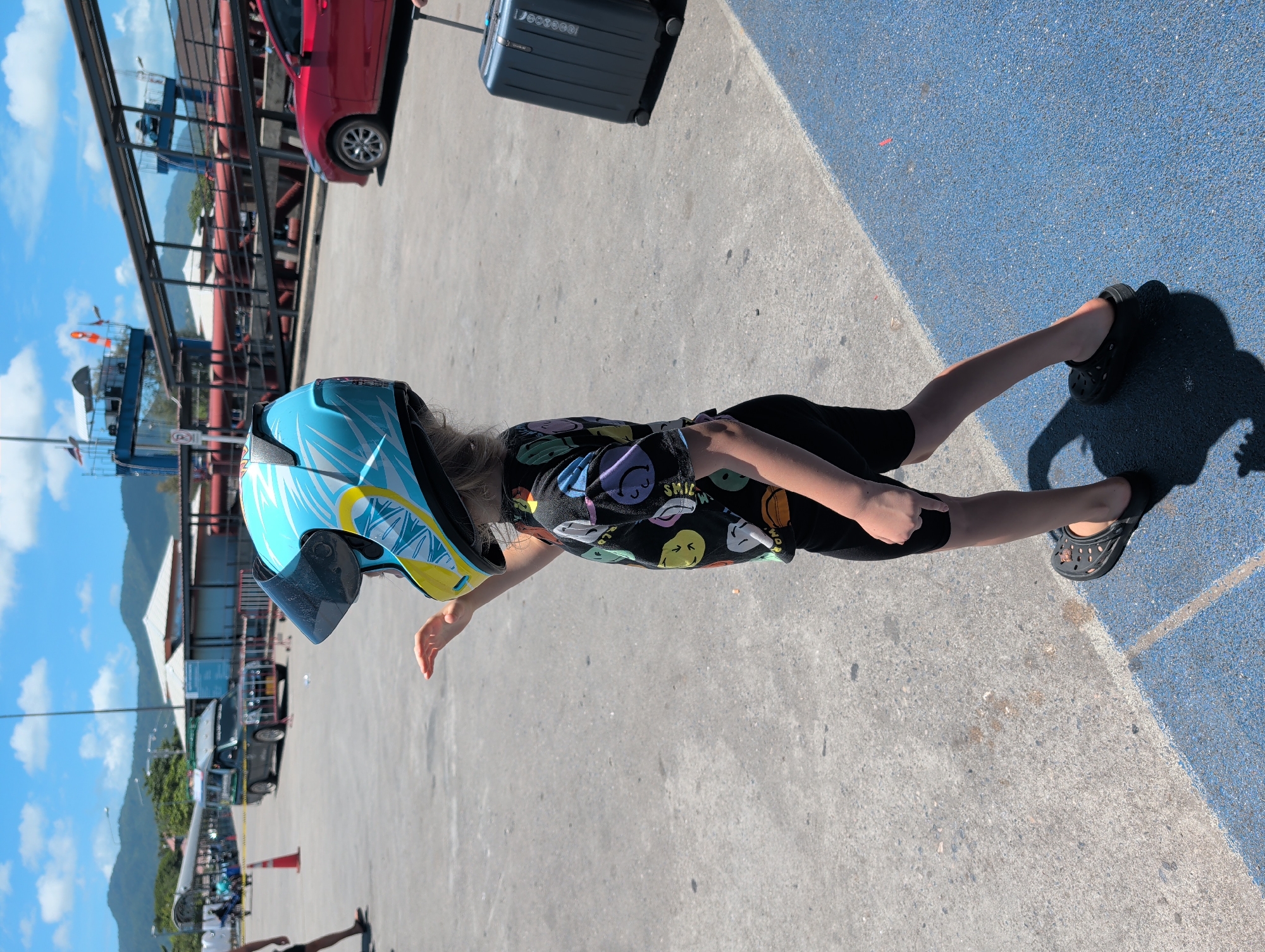 A child wearing a blue motorcycle helmet and black Crocs is standing on a concrete surface with blue painted sections, adjacent to a luggage case and red car. In the background is a coastal landscape with buildings, boats, and a pier-like structure, under a blue sky with scattered clouds. The image was taken in ตำบลเกาะพะงัน, Thailand.