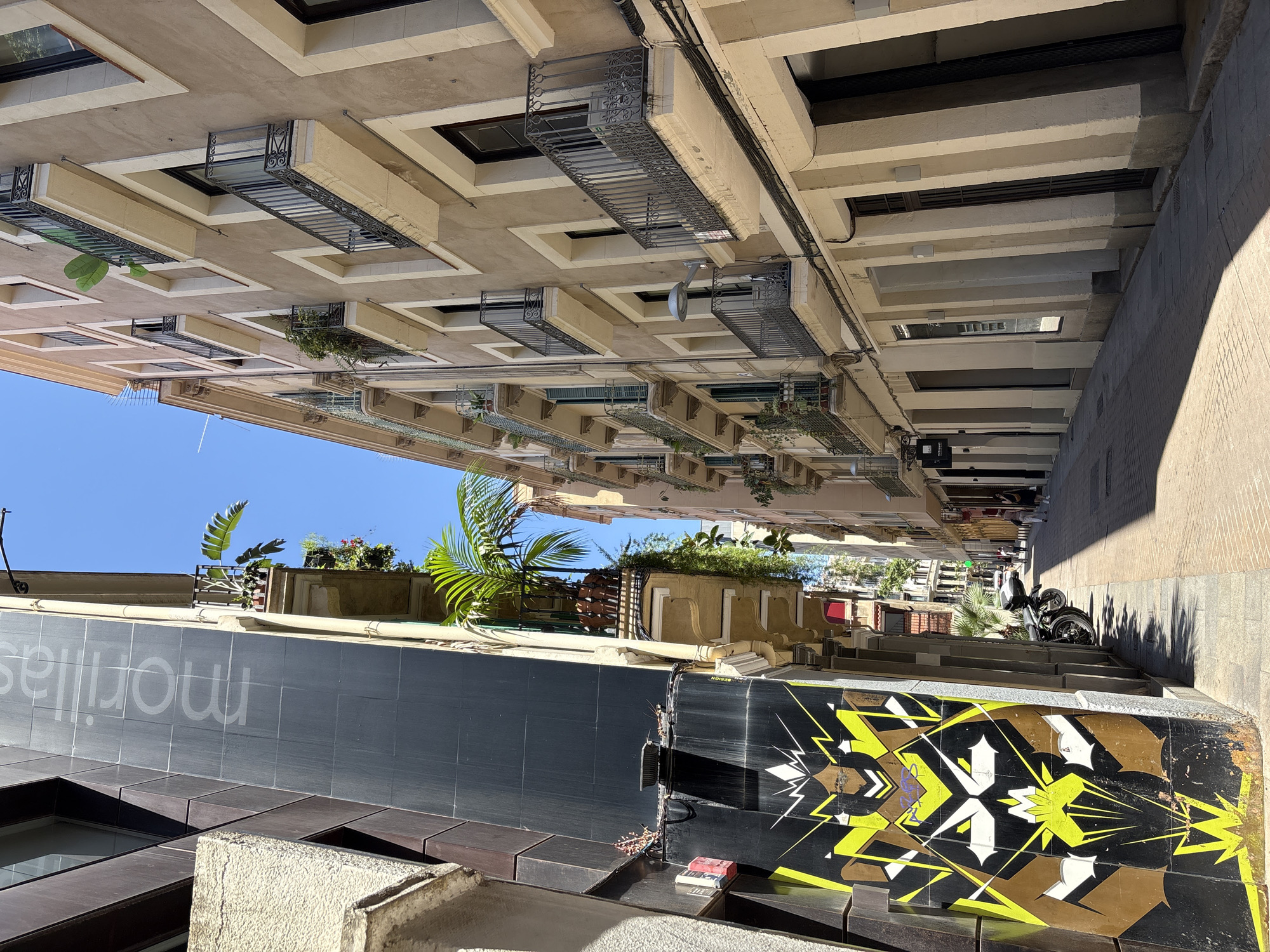 This image captures a narrow street in Barcelona, Spain, flanked by tall buildings on either side. The architecture suggests a Mediterranean style, with weathered facades and wrought-iron balconies adorned with potted plants, including a prominent palm tree. The sunlight casts sharp shadows, indicating a bright, sunny day. On the left, a dark-colored building features a large, striking mural in black, yellow, and white, with a vertical text that appears to read "morillas" in a lighter tone. The right side of the street is lined with buildings exhibiting a lighter, sandy-colored facade, with multiple floors featuring balconies. A few people are visible in the distance, walking along the street, and a motorcycle is parked on the left side, near the mural. The street is paved with interlocking bricks, and the sky is a clear, vibrant blue.