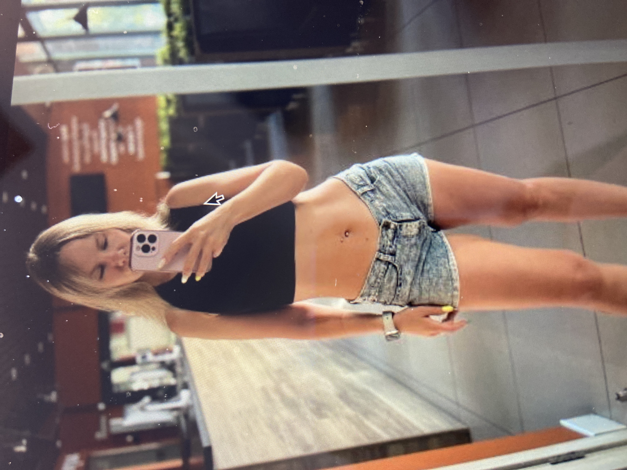 A blonde woman stands in a gym, taking a mirror selfie with her phone. She is wearing a black crop top and denim shorts. Her hair is down, and she is wearing a watch on her left wrist. The gym appears to be well-lit and modern, with exercise equipment visible in the background. The overall impression is that of a fitness enthusiast capturing a moment after a workout.