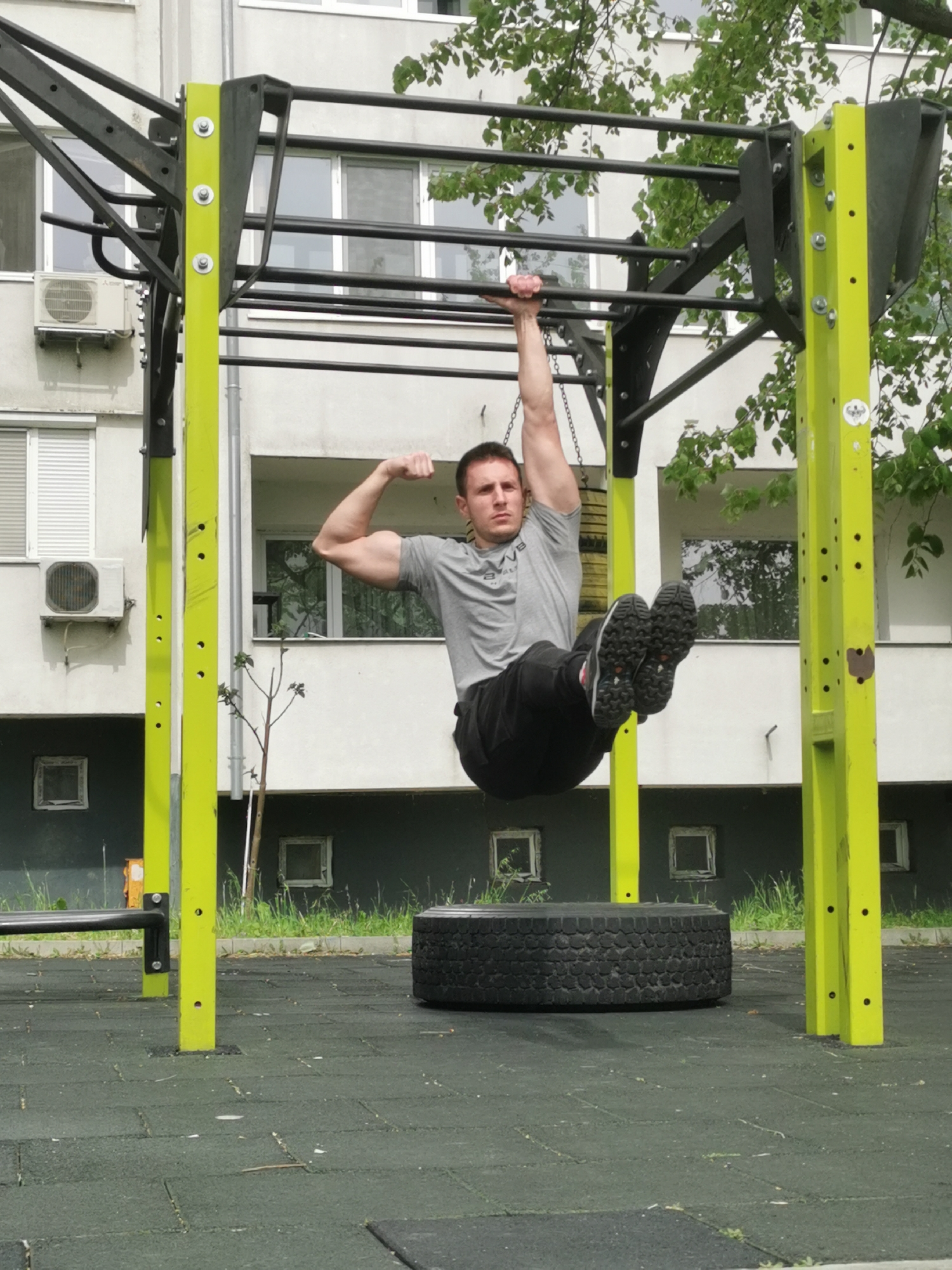A man is suspended from a pull-up bar in what appears to be an outdoor exercise area. He is hanging with one arm while flexing the bicep of the other arm. The pull-up bar is part of a larger metal structure with yellow vertical supports. Behind the man is a multi-story building with windows and air conditioning units. A large tire sits on the ground beneath him, resting on dark green rubber flooring. The exercise area is located in the city of Burgas, Bulgaria.