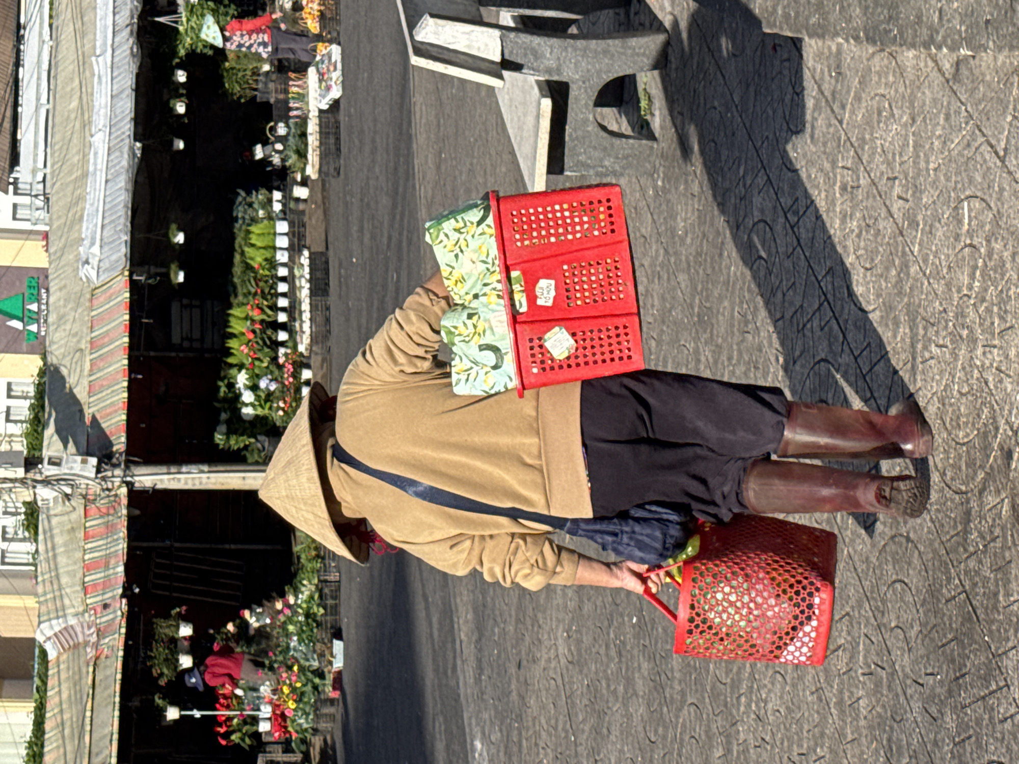 The image depicts a person walking away from the camera, carrying two red baskets. The person is wearing a brown hoodie, black pants, and brown boots, and a conical hat. The left basket is filled with red items, possibly fruit or vegetables. The right basket contains rolled items covered in fabric with a green and yellow pattern, and a white label reading "FUJI TO HUU". In the background, a market stall selling plants and flowers is visible, with awnings and various potted plants. A stone bench is situated to the right of the person. The scene appears to be an outdoor market in Da Lat, Vietnam, characterized by the conical hat, the style of the market stalls, and the presence of plants and flowers typical of the region. The ground is paved with patterned tiles.