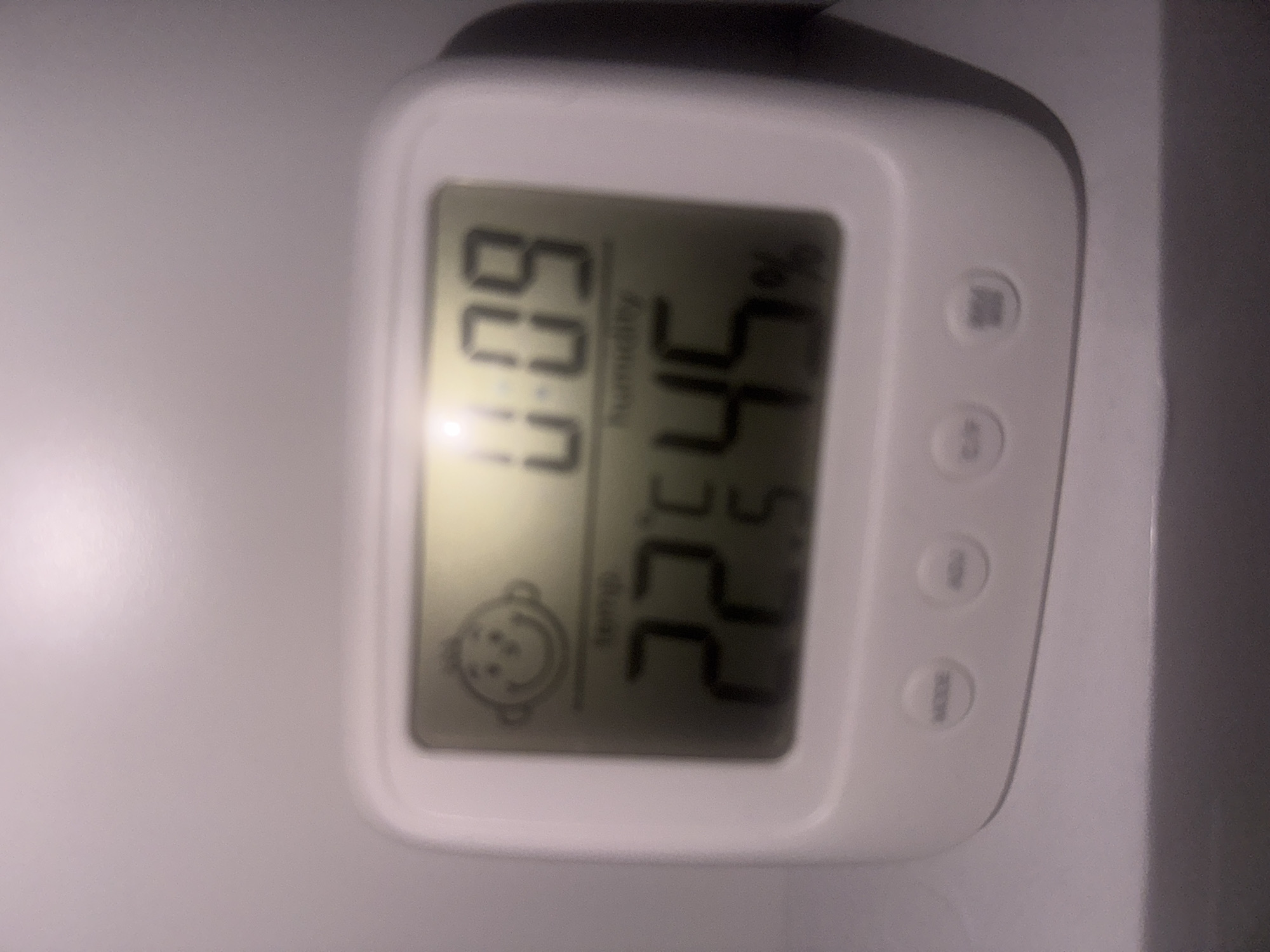 The image is a close-up, slightly out-of-focus photograph of a white digital thermometer and hygrometer, likely a baby monitor. The device displays the time as 11:09, temperature as 22.5 degrees Celsius, and humidity as 45%. A small, simplified graphic of a smiling baby's face is visible above the temperature reading. Below the display are several circular buttons labeled "MODE", "ADJ", and "CH/MAX/MIN". The device is resting on a white surface, possibly a shelf or table, with a soft, diffused light source creating a slight glow from above. The background is a plain, light-colored surface. The image does not contain information about a specific city or location.