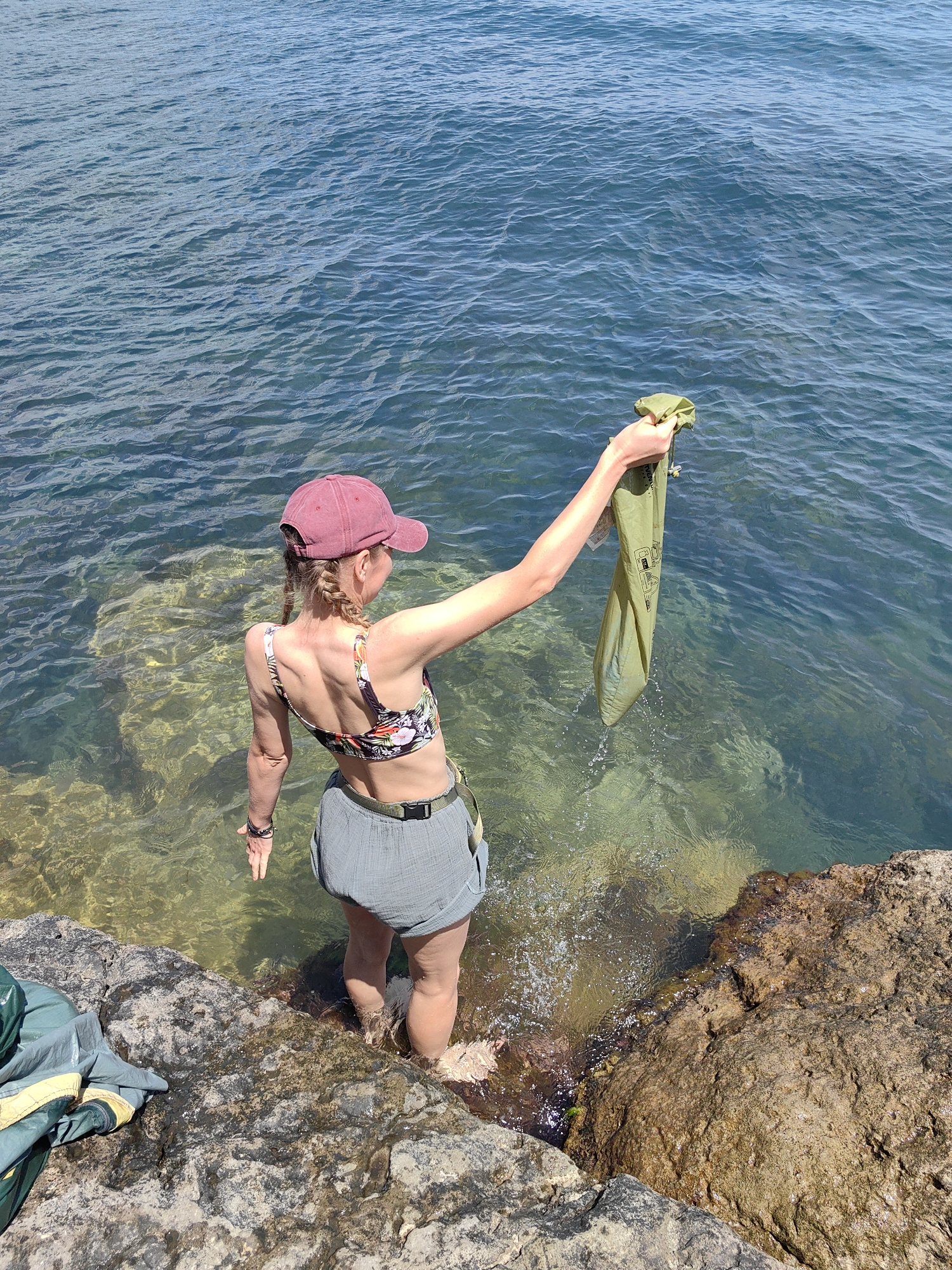 A woman wearing a burgundy baseball cap, a floral print bikini top, and grey shorts stands at the edge of rocky shoreline, with her feet submerged in clear, shallow water. She is holding a green fabric bag aloft in her right hand, with water dripping from it. The background is the blue sea, with visible ripples and sunlight reflecting on the surface. The shoreline is composed of large, weathered rocks.