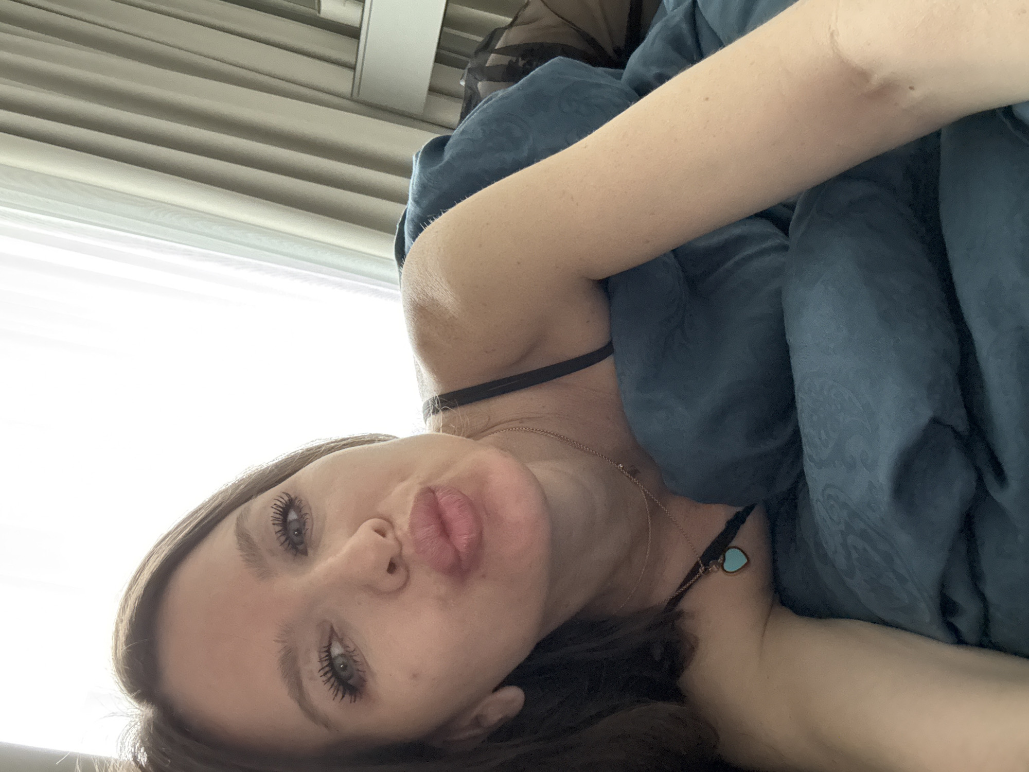 A woman is lying down, partially covered by a blue patterned blanket. She has brown hair and visible makeup including mascara. She is wearing a black strappy top, a thin gold necklace, and a necklace with a small heart-shaped blue pendant. The background shows white blinds. No information in the provided image confirms that the location is in the city of Khimki, Russia.