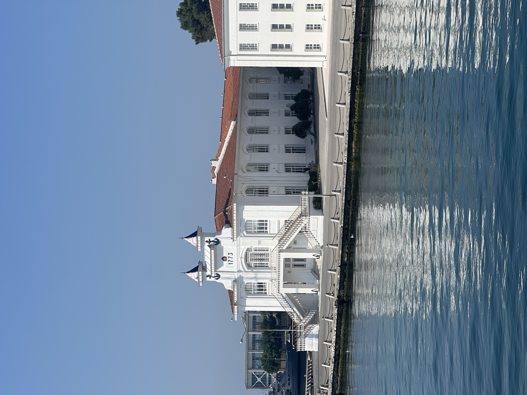 The image depicts the Heybeliada Naval High School building in Istanbul's Prince Islands district. The white, colonial-style structure features a prominent central entrance with a grand staircase, flanked by two smaller towers topped with spires. The year "1773" is visible above the entrance, along with anchor motifs. The building has a red-tiled roof and numerous arched windows. To the right, another white building with a similar red roof is partially visible. The structures are situated along a waterfront promenade, separated from the blue water by a stone barrier and a chain fence. The water reflects the sunlight and the buildings, creating a shimmering effect. In the background, a large modern building with a glass facade and a navy vessel are visible. The sky is clear and blue, indicating a sunny day. No people are clearly discernible in the image.