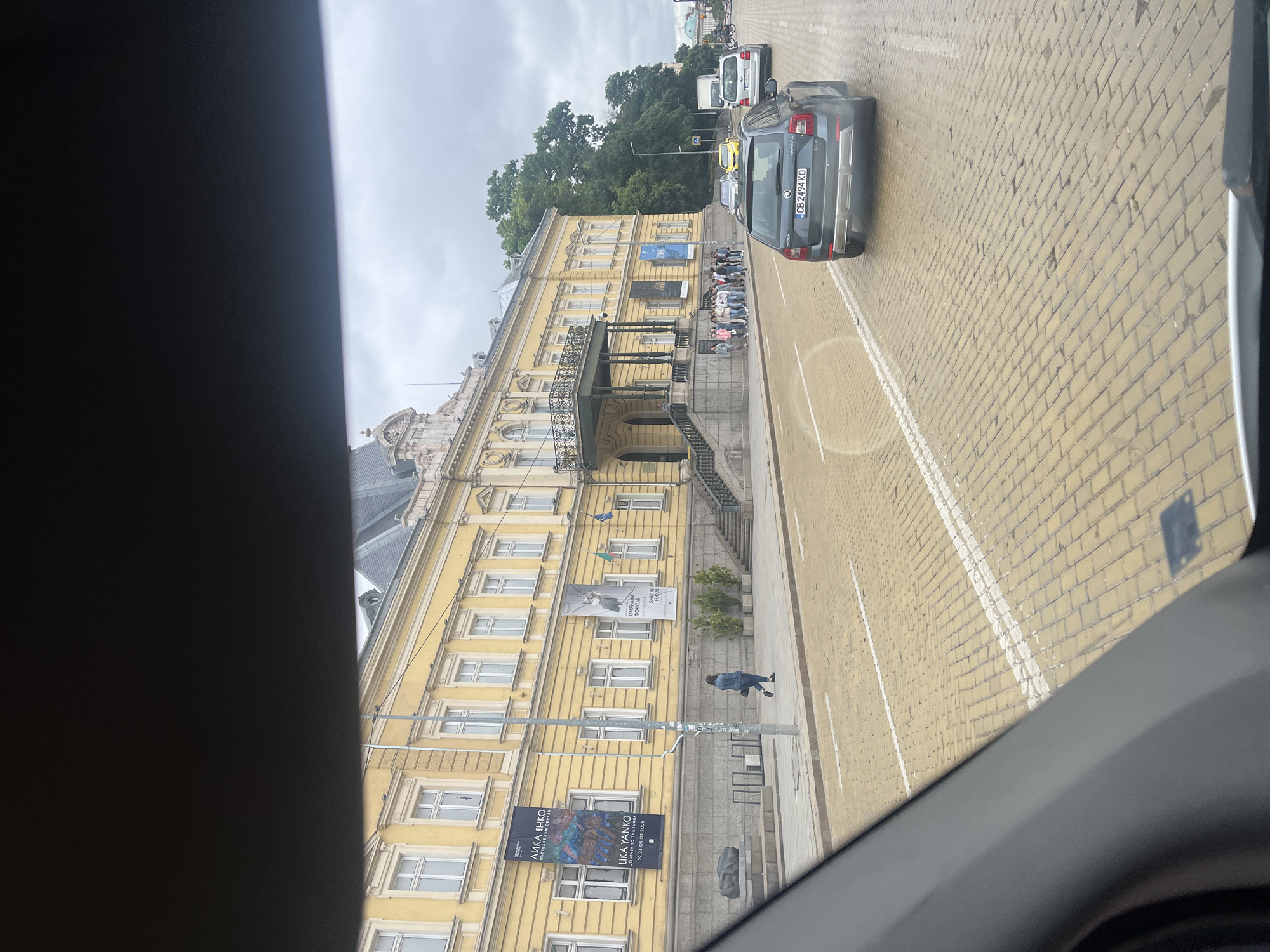 The image shows a yellow building with a grand entrance and balconies, possibly a museum or public building, with a street in front of it paved with yellow bricks. Several cars are visible on the street, including a grey Skoda Octavia with license plate CB 2494 KO. A group of people are standing on the sidewalk in front of the building, and a single person is walking away from the camera on the sidewalk. Several banners are hanging on the building, one of which advertises "LIKA YANKO JOURNEY TO THE IMAGE." The sky is overcast.