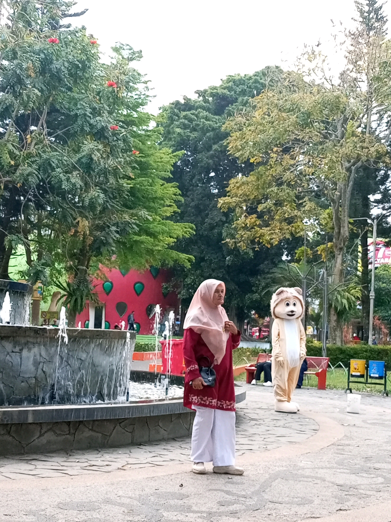 This image depicts a scene in an outdoor park setting, potentially within the city of Batu, Indonesia, indicated by the style of vegetation and general atmosphere. In the foreground, a woman wearing a pink hijab and a dark red outfit with white embroidery stands in profile, looking to her left. She has a dark crossbody bag. To her right, a person in a tan mascot costume resembling a cartoon lion stands facing forward. The mascot has large eyes and a friendly expression.
Behind the woman, a multi-tiered stone fountain with water cascading down is visible on the left. Beyond the fountain and mascot, a bright red, strawberry-shaped building with green accents serves as a whimsical structure, possibly an attraction. Lush green trees with some showing red berries, and other foliage fill the background. The ground is paved with stone and concrete, with some fallen leaves. In the mid-ground, there are red benches and trash receptacles. The lighting suggests it is daytime, possibly overcast.