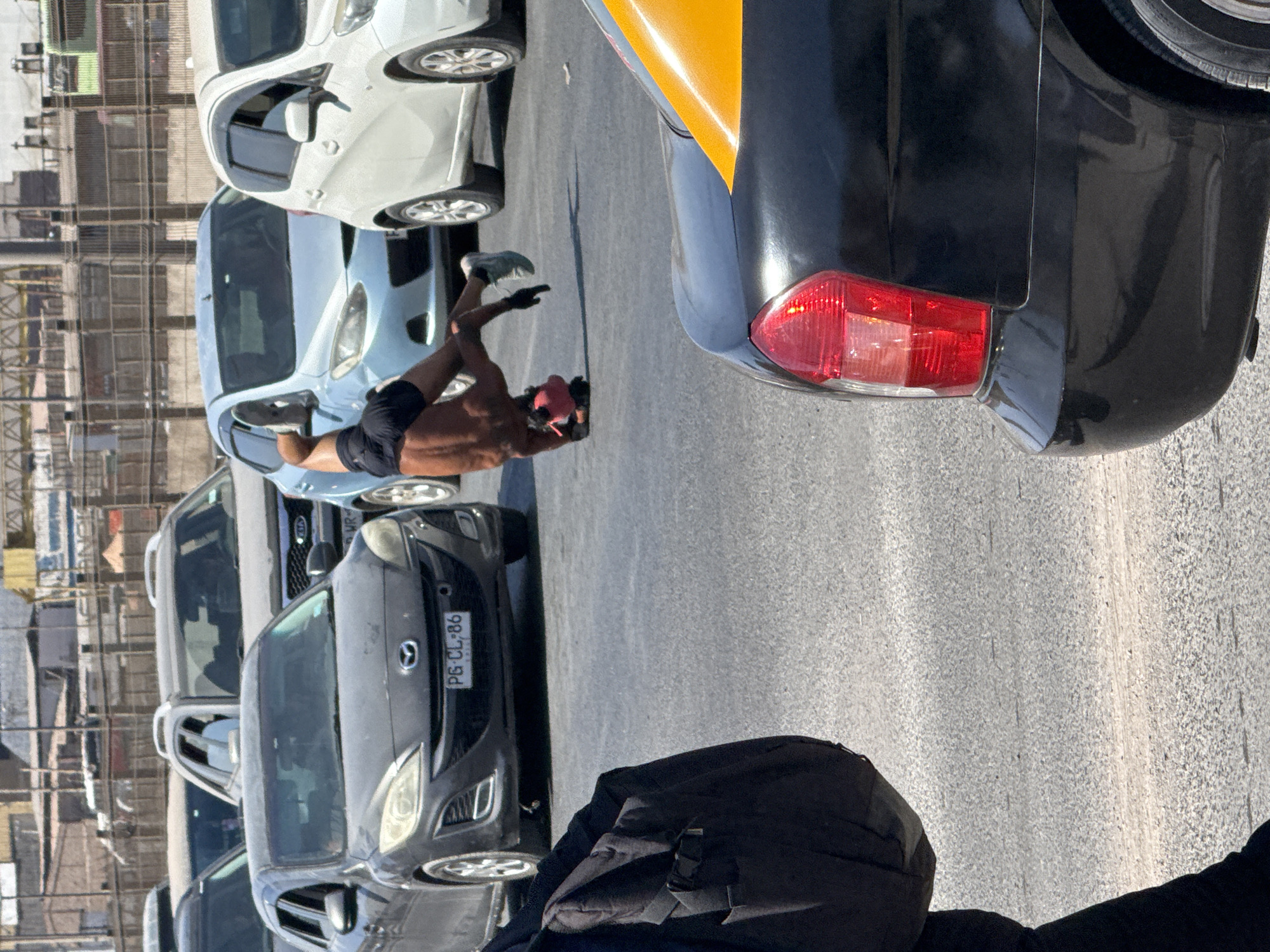 The image depicts an urban street scene under bright daylight in Unknown, Unknown, featuring a street performer amidst stopped traffic. In the center, a shirtless individual with dark shorts and light-colored sneakers is performing an inverted balance, possibly a handstand or headstand, on a red object on the asphalt pavement. They are positioned between several stationary vehicles.
To the performer's immediate left, a dark grey Mazda sedan with license plate "PG CL:86" is stopped. Behind the performer, a light blue sedan and a white hatchback are also stationary. Further back, a grey Kia SUV with roof rails and other vehicles are visible, indicating a traffic jam. In the immediate foreground on the right, the rear portion of a black sedan with a prominent red taillight and a partial yellow stripe on the trunk is visible, suggesting a taxi. On the far left foreground, the lower torso and left leg of a person wearing dark clothing and carrying a dark backpack are partially visible, walking away from the camera. The background consists of distant urban structures, fences, and utility elements under a clear sky. The road surface is dry asphalt.
