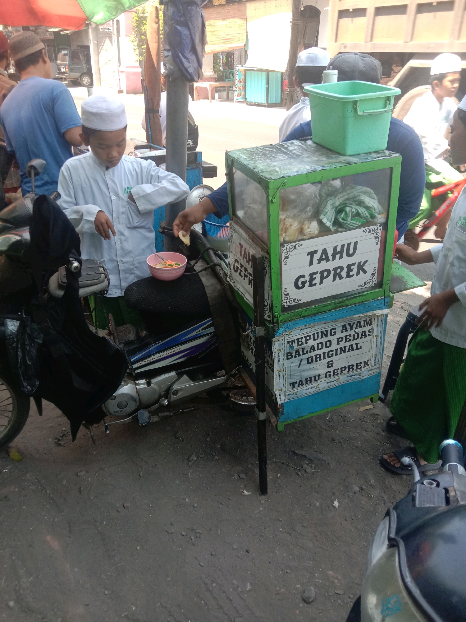 The image depicts an outdoor scene in the city of Karang Anyar, Indonesia. A young boy wearing a white cap and a white shirt is standing behind a mobile food cart. He appears to be interacting with the food cart, with one hand near a pink bowl containing food. The food cart is decorated with signs advertising "TAHU GEPREK" and "TEpung Ayam Balado Pedas / Original & Tahu Geprek." A green plastic container is placed on top of the food cart. Beside the cart, an adult wearing a green sarong and sandals is visible. In the background, other people, a motorcycle, and various stalls or structures are present. The ground is a dirt or gravel surface.