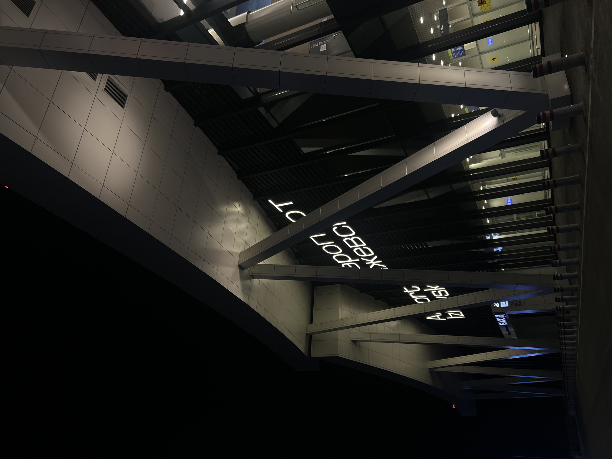 The image captures the interior or immediate exterior architecture of a modern building at night or in a dimly lit environment, seen from a low angle looking upwards. The structure is characterized by prominent, light-colored, angular beams that intersect and crisscross, forming a complex geometric pattern against a dark background. These beams appear to be part of the building's support system or a decorative architectural feature.
The walls are clad in light grey or white panels, creating a tiled effect with visible seams. Some sections of the ceiling or upper structures are dark, possibly revealing exposed utilities or dark soffits. Numerous artificial lights illuminate the scene; some are embedded in surfaces, while others shine from behind windows or translucent panels in the background, suggesting further illuminated spaces beyond. Several metal grates or vents are visible on the wall surfaces. Along the right side of the image, multiple cylindrical elements, possibly support pillars or pipes, with distinct red and white banding, extend upwards.
No people are visible, and no specific activity is taking place within the frame, emphasizing the architectural elements. The time of day is clearly night, or it's a very dark indoor space, given the reliance on artificial lighting.
Several pieces of glowing white text, primarily in Cyrillic, are displayed on the main beams, acting as a prominent feature. Clearly visible words include "КЕМЕРОВО" (Kemerovo), "НОВОКУЗНЕЦК" (Novokuznetsk), and "АЭРОПОРТ" (Airport), strongly suggesting the location is an airport, specifically Kemerovo International Airport (or Novokuznetsk, as both city names are present). Another smaller sign in the background shows "ВХОД" (Entrance) and yellow arrow symbols, common airport signage. The architecture is sleek and modern, fitting with a contemporary transport hub. The location context provided confirms this is in Russia.