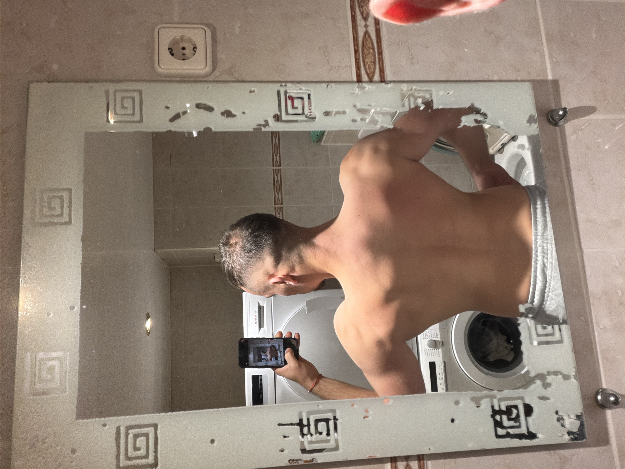 A shirtless male is standing in a bathroom in an apartment, taking a picture of himself in a mirror with a smartphone. The mirror is decorative and appears to have some damage. Reflected in the mirror are the man, a white washing machine, and bathroom wall tiles. A power outlet is visible on the wall beside the mirror. The location is possibly Lelystad, Netherlands.