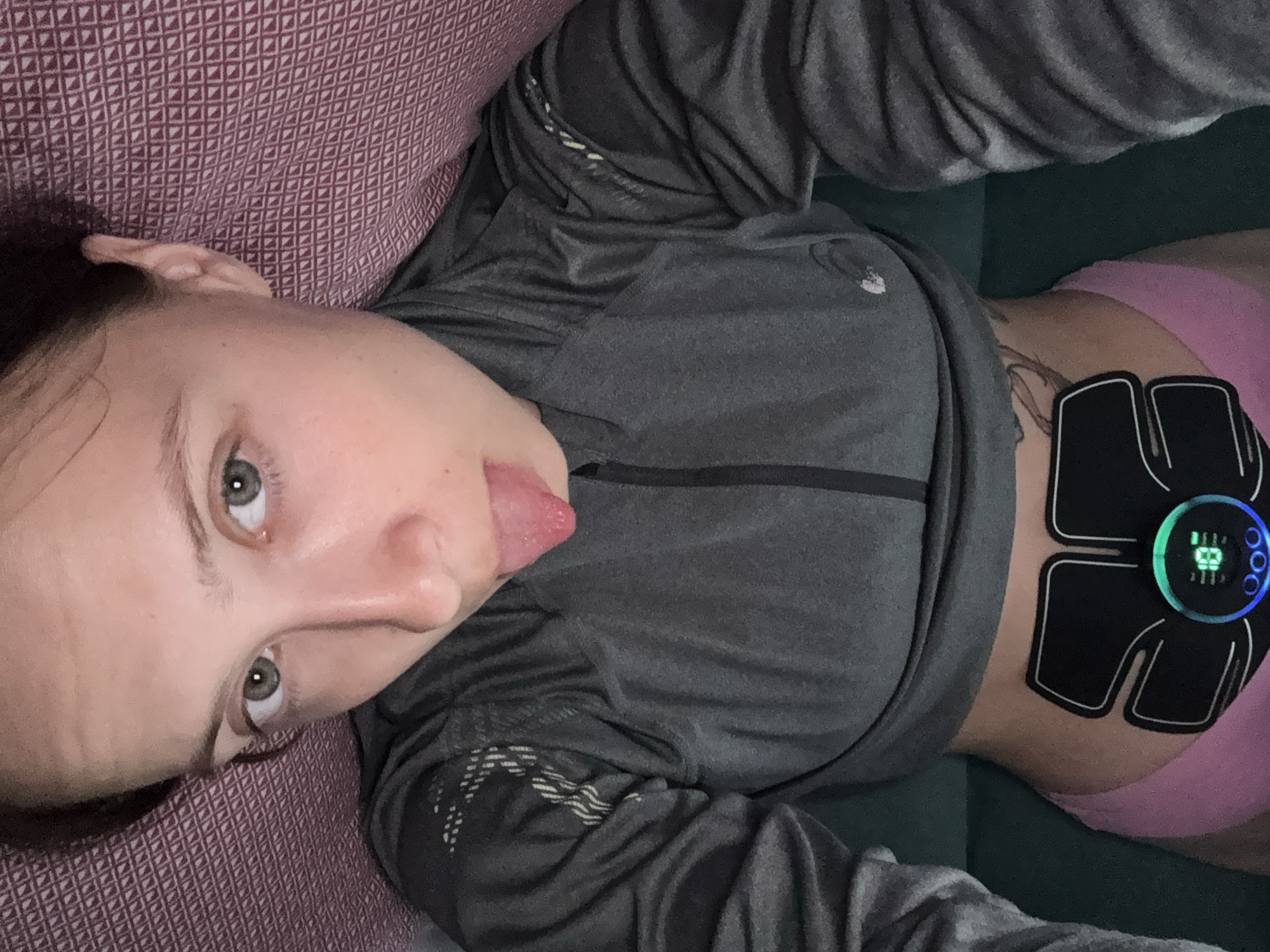 An overhead shot captures a light-skinned individual, likely female, with dark hair and grey-green eyes, lying in an indoor setting in Прикамский, Russia. She wears a dark grey, zippered athletic top with patterned sleeves and pink underwear, exposing her abdomen. An active black electronic muscle stimulator (EMS) device is affixed to her bare midriff, displaying "18" with blue and green illumination on its central circular control unit. The individual looks directly into the camera, extending her tongue. The immediate background behind her head is a textured, reddish-purple fabric, while a dark teal-green surface is visible to her sides.