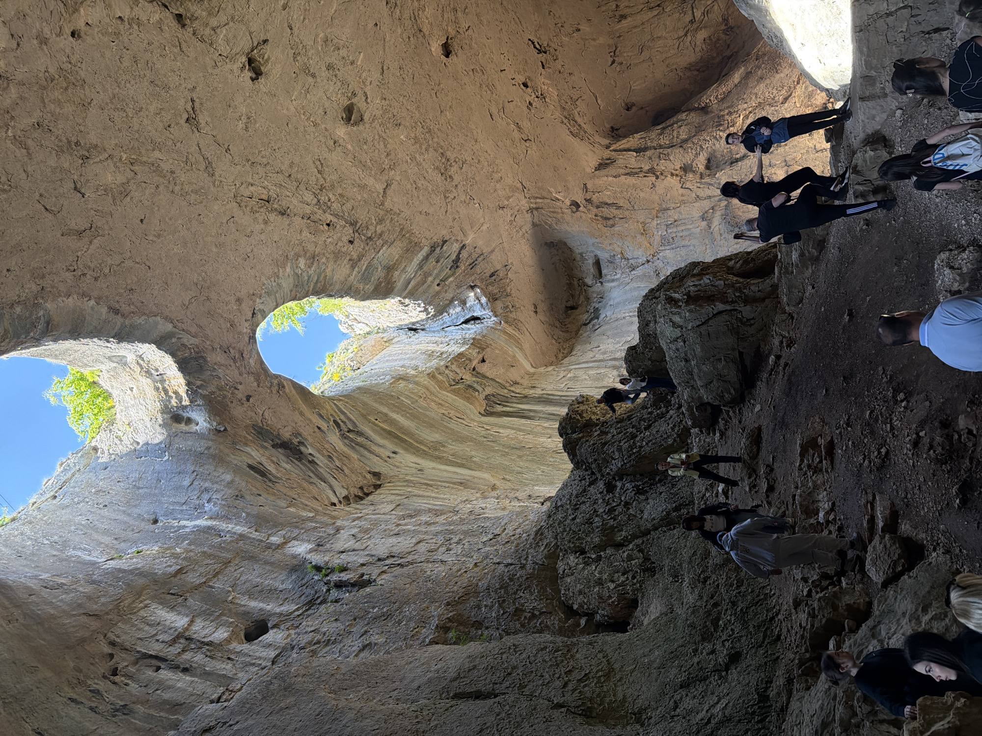 This is a low-angle, wide-shot photograph of the Devetashka Cave near Karlukovo, Bulgaria. The focus is on the cave's interior, emphasizing its geological features and the people exploring it. The cave's walls are predominantly light-colored rock with visible striations and formations, including two large circular openings allowing daylight to penetrate. Patches of green vegetation cling to the cave openings where the sky is visible. A group of people are visible along the rock face to the right. Some are standing and looking up into the cave, and others are assisting each other in climbing over the rocks. They wear a variety of casual clothing.