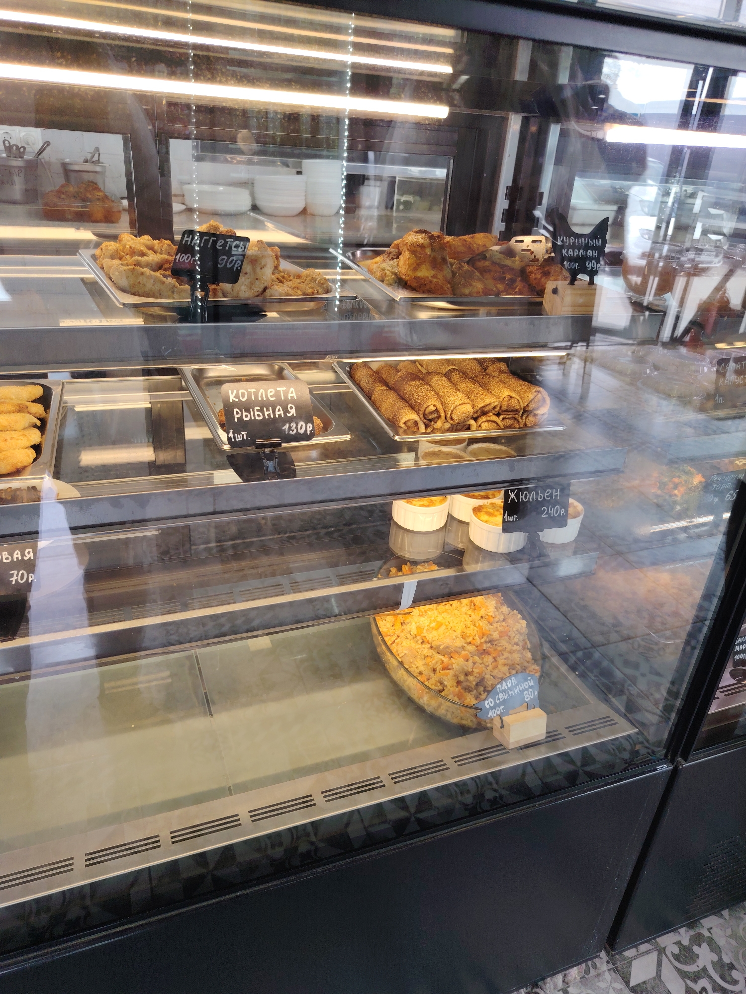 The image shows a display case in a food establishment. The display case is divided into multiple shelves, showcasing various food items. On the top shelf, there are what appear to be fried chicken pieces, with a price tag indicating "100р 90р" and the text "НАГГЕТСЫ". To the right of this, there are more fried chicken items with a tag featuring a chicken silhouette and the text "КУРИНЫЙ КАРМАН 100г. 90р.".
The middle shelf displays several trays of food. On the left, there are golden-brown breaded items, possibly fish or chicken. In the center, there's a tray of rolled crepes or blinis, and a price tag reads "КОТЛЕТА РЫБНАЯ 1 шт. 130р.". To the right of the crepes, there are small ramekins filled with a baked dish, with a price tag stating "Жюльен 1 шт. 240р.". Further to the right, there are other food items in containers, one with visible green herbs.
The bottom shelf contains a large glass bowl filled with what appears to be pilaf or rice with vegetables, and a price tag reading "ПЛОВ СО СВИНИНОЙ 100г. 80р.". The background shows reflections of lights and possibly other parts of the establishment. The flooring visible at the bottom of the image has a patterned tile design. The establishment is located in Kagal'nitskaya, Russia. There are no people visible in the image. The food items are arranged in trays and containers within the refrigerated display case.