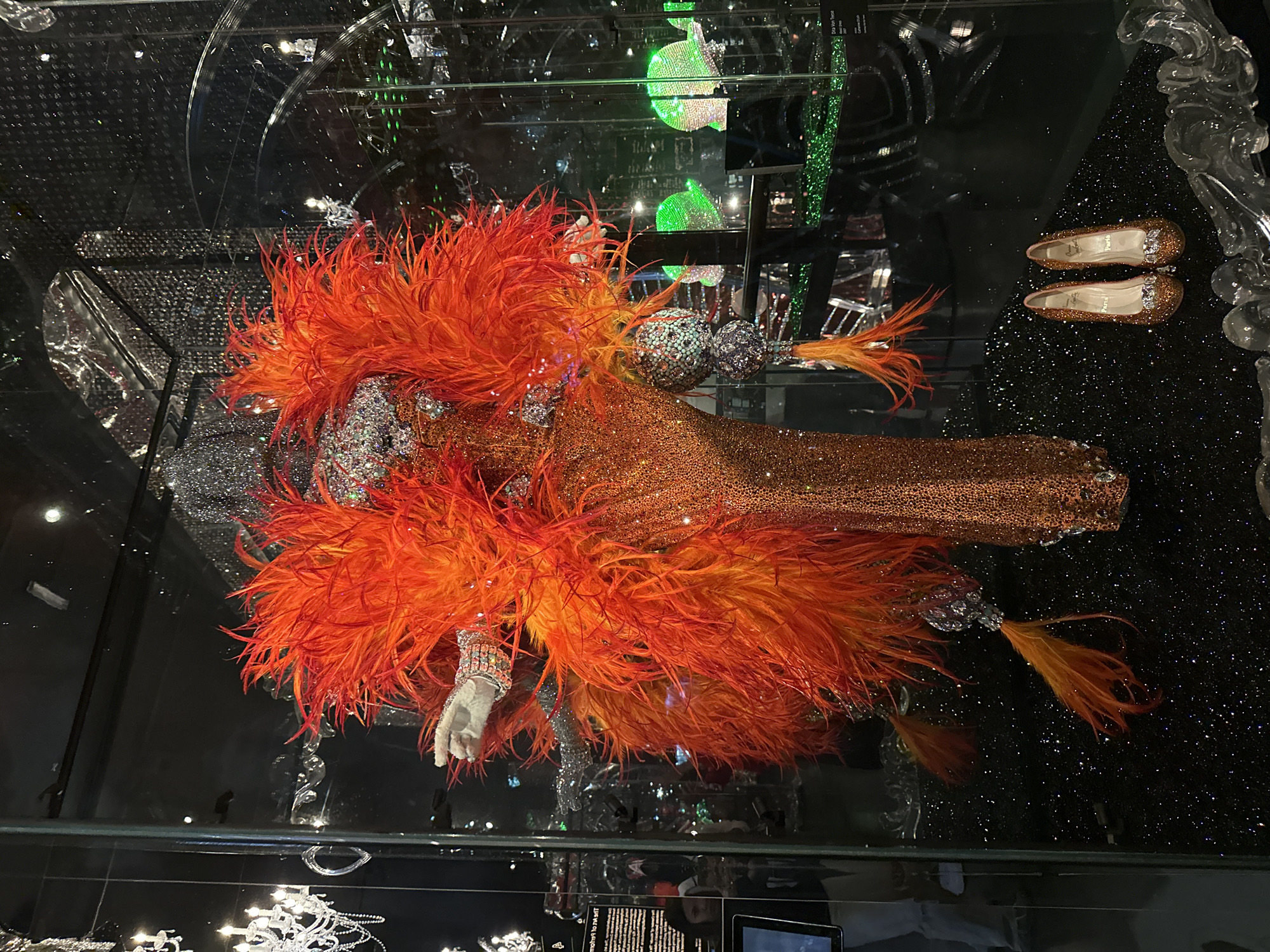 A dazzling display is presented within a glass enclosure at the Swarovski Kristallwelten in Wattens, Austria. The focal point is a mannequin adorned in an elaborate, shimmering orange gown, encrusted with countless crystals that catch the light. The gown features a dramatic, voluminous collar of bright orange and red feathers that extend outwards, framing the upper body. Two bejeweled, spherical ornaments hang from the sides of the mannequin, also sparkling with crystals. A single white-gloved hand, accented with a crystal bracelet, is extended from the mannequin's left side. At the base of the display, on a black, glitter-covered surface, rests a pair of small, sparkling orange ballet flats, each embellished with a crystal bow and the word "Paris" inscribed on the sole. The background of the enclosure is a dark, reflective surface, with hints of other crystal displays and lighting visible, including a chandelier on the left and other green crystal objects further back. Two figures are partially visible through the glass on the left side of the enclosure, one appearing to be a woman looking at an information plaque.