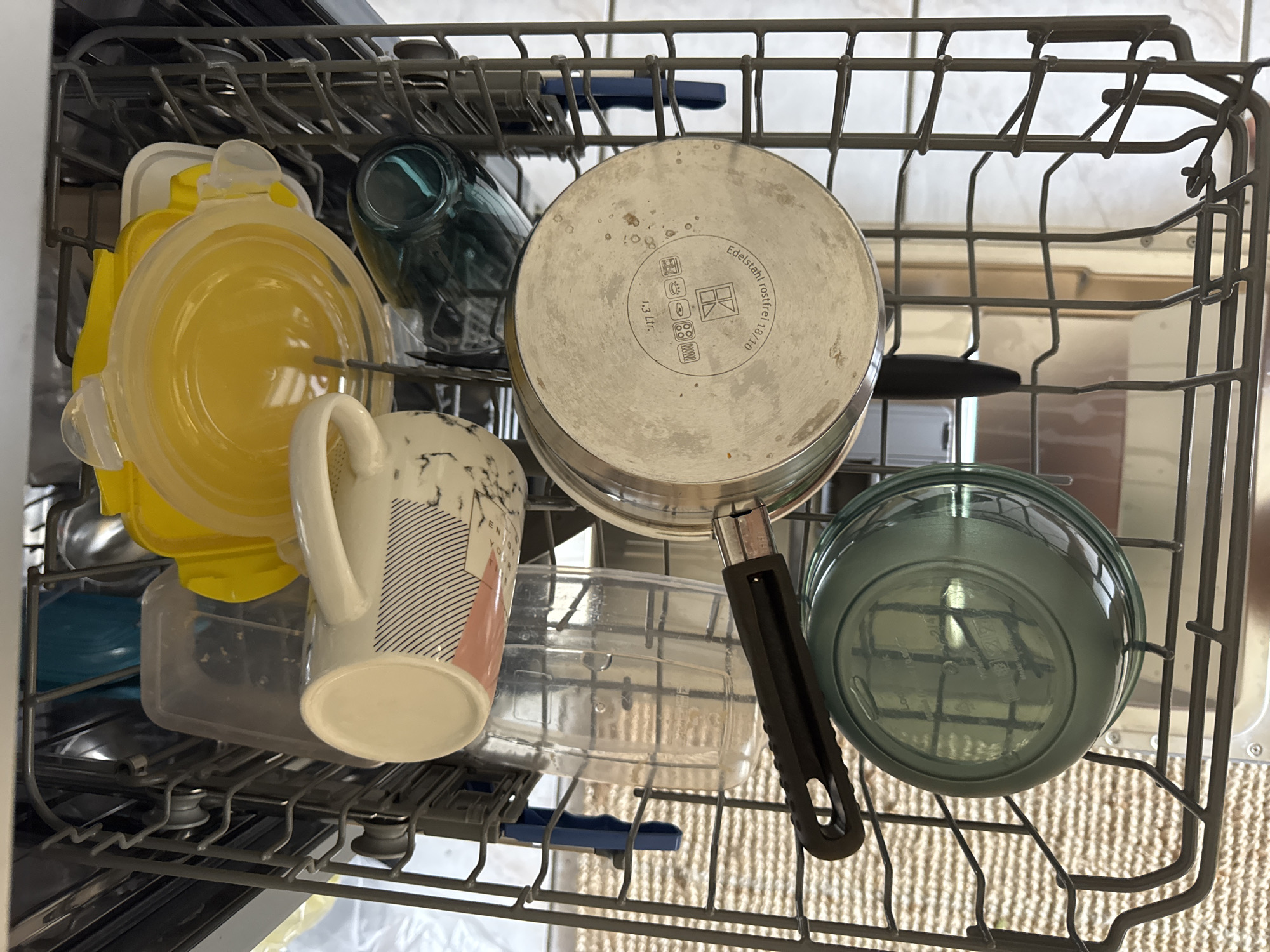 The image shows the interior of a dishwasher in Höhenkirchen-Siegertsbrunn, Germany. The dishwasher rack is filled with various kitchen items, including a yellow lidded container, a patterned mug, a clear plastic measuring cup, a dark green lidded container, and a stainless steel pot with a black handle. The items are arranged in a seemingly organized manner for washing. The background reveals the metallic interior of the dishwasher.