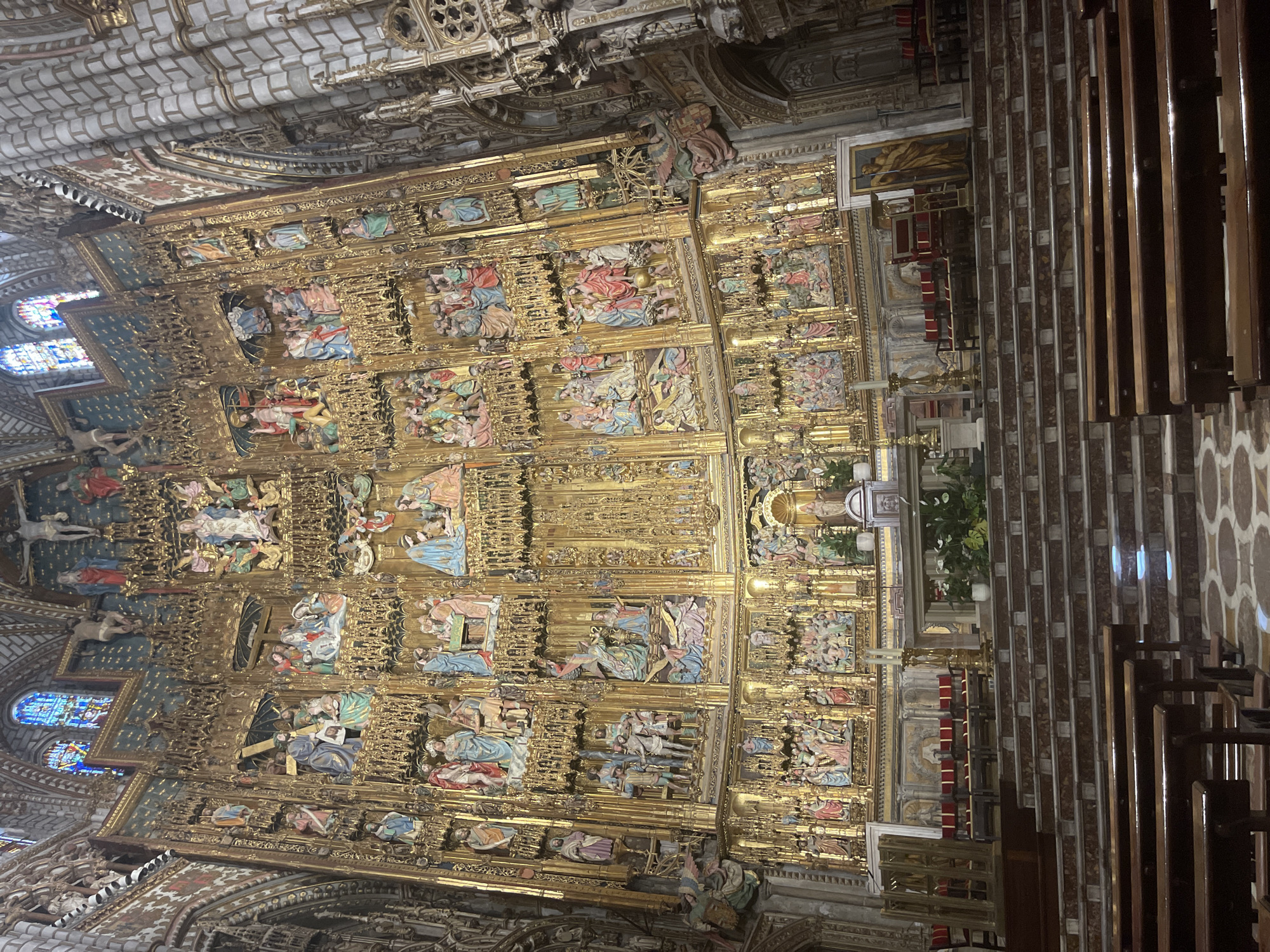 This image depicts the elaborate golden reredos behind the altar of the Cathedral of Toledo in Toledo, Spain. The reredos is a massive, multi-tiered altarpiece intricately carved and gilded, featuring numerous polychrome religious figures and scenes from the life of Christ and the Virgin Mary. The figures are arranged in rows and niches, creating a densely ornamented spectacle. In the foreground, wooden pews are visible, leading up a marble staircase towards the altar area. The architecture of the cathedral, with its stone columns and stained-glass windows in the upper part of the image, provides a grand and solemn backdrop. The lighting suggests an interior scene, likely illuminated by natural light filtering through the windows and possibly artificial lighting.