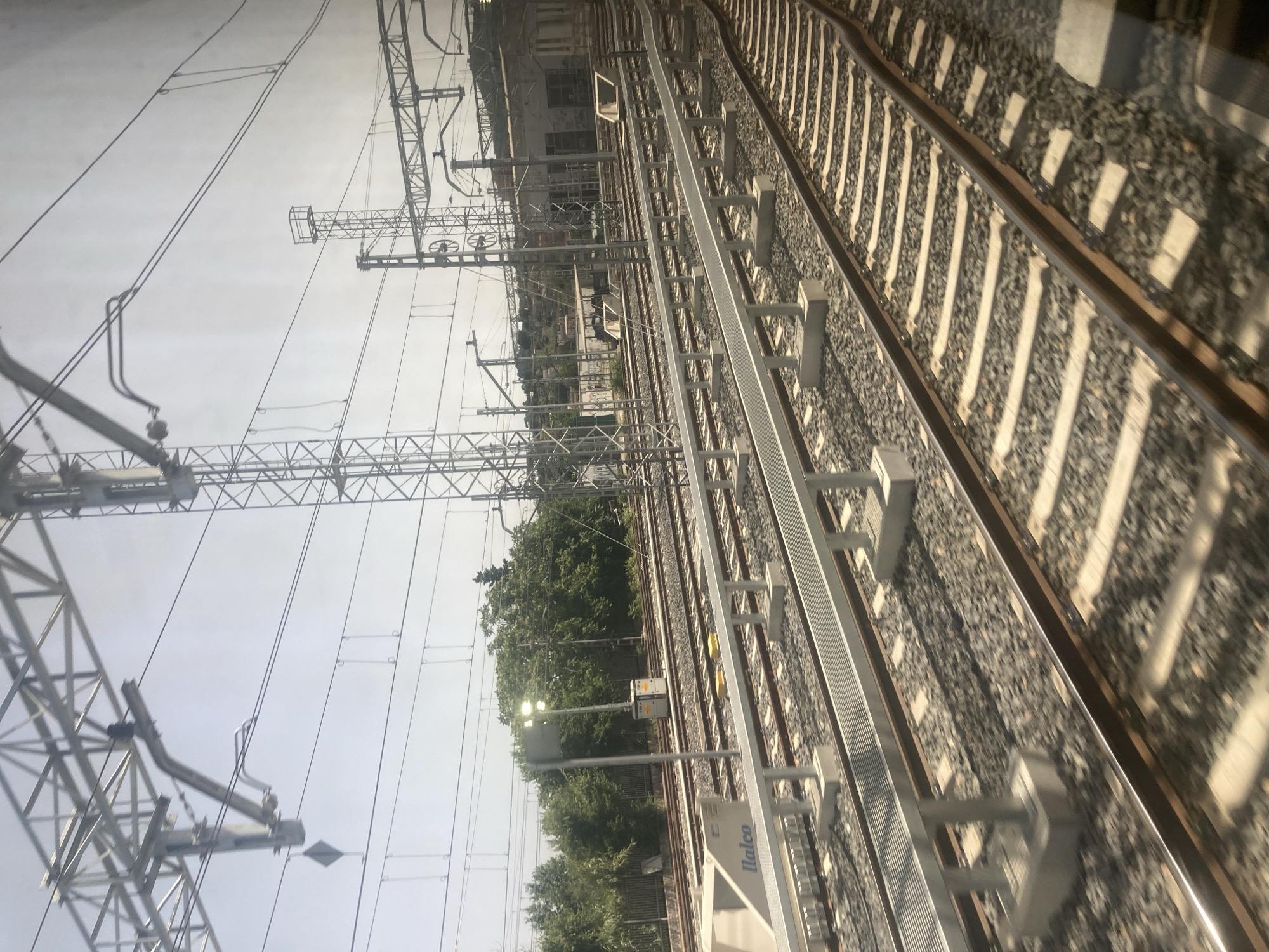 The image depicts an outdoor view of a train station with multiple sets of railway tracks in Montcada i Reixac, Spain. The scene is dominated by the infrastructure of the railway system, including overhead power lines supported by tall metal pylons, and metal safety barriers running alongside some of the tracks. The tracks themselves are made of gravel and sleepers, with steel rails. In the foreground, a section of a modern platform or walkway with a metal grating surface is visible. To the left, a white structure with the word "llaleco" printed on it is present. In the mid-ground, there is a lamppost with several lights, and beyond that, a dense line of trees forms a green backdrop. Further in the background, there is a low-rise building with windows and a hilly landscape with scattered houses. The sky is bright and hazy, suggesting daylight. There are no people visible in the image.