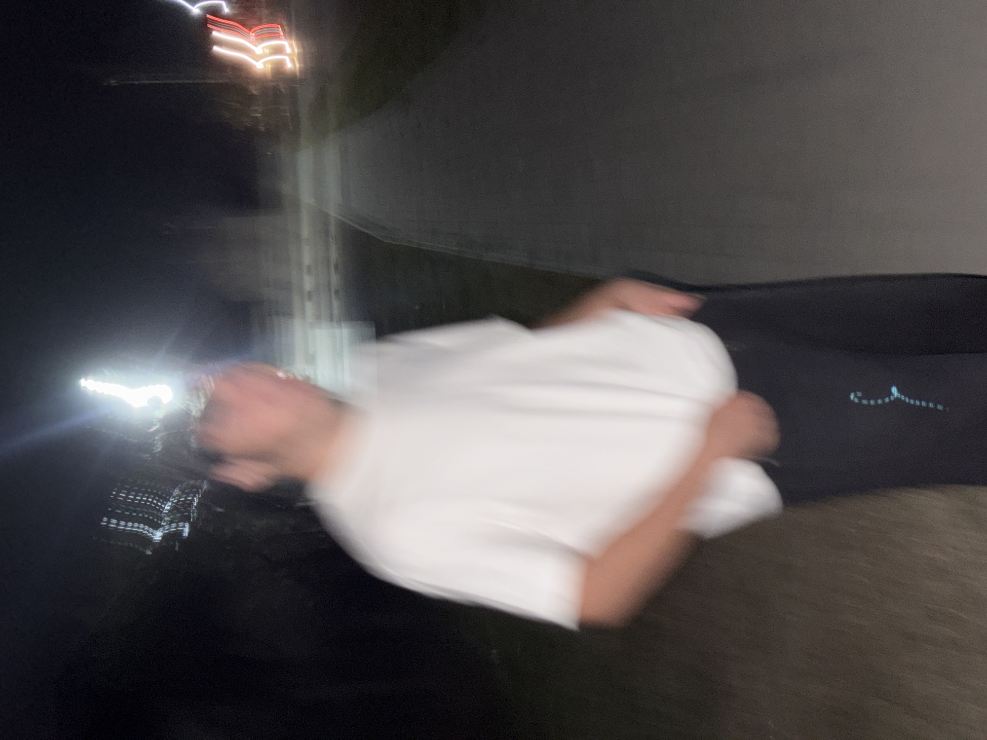 The image is a blurry, low-light, nighttime photograph of a person standing on a paved walkway next to a grassy area. The person is wearing a white t-shirt and dark pants, with their hands in their pockets. In the background, there are lights from what appears to be a street or a building. The location is identified as Илсхан-Юрт, Russia. The overall impression is one of motion blur, possibly due to a long exposure or movement during the photograph.