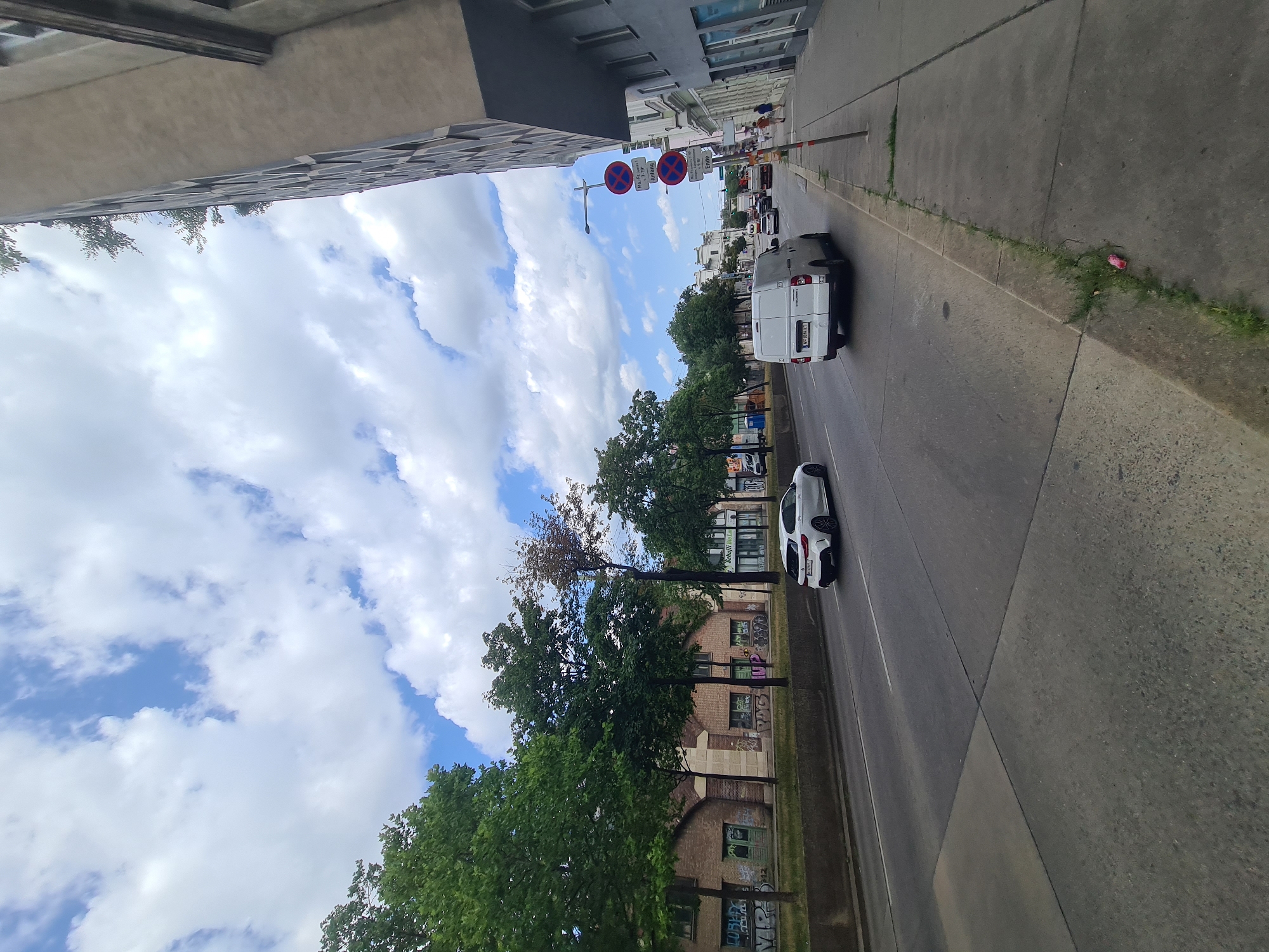 The image shows a street scene in Vienna, Austria, on a cloudy day. The perspective is from street level, looking down a wide road with two lanes of traffic in each direction. On the left side of the road, there are trees and a brick building with graffiti. On the right side, there is a sidewalk, a tall modern building, and some older, multi-story buildings. Traffic includes a white car, a white van, and other vehicles in the distance. Several traffic signs are visible on the right side of the street, including parking restrictions. Two people are walking on the sidewalk on the right. The sky is filled with white, fluffy clouds.