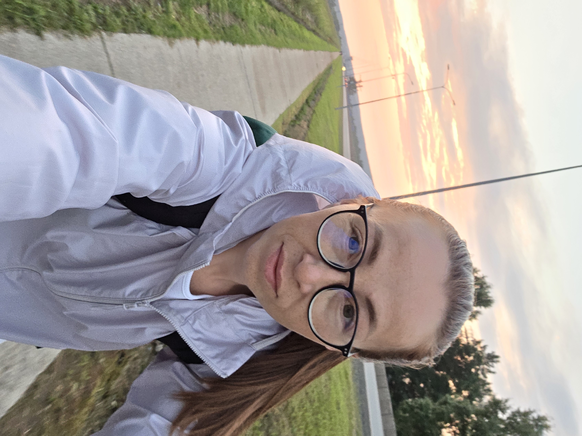 A woman wearing glasses and a light-colored hooded jacket is taking a selfie outdoors during sunset. She has long, brown hair pulled back into a ponytail and is wearing a white tank top underneath her jacket. The background shows a paved path lined with grass, a road with streetlights, and trees. The sky is filled with soft, pastel colors from the sunset. The woman appears to be walking or cycling on a path in the city of Chaikovsky, Russia.