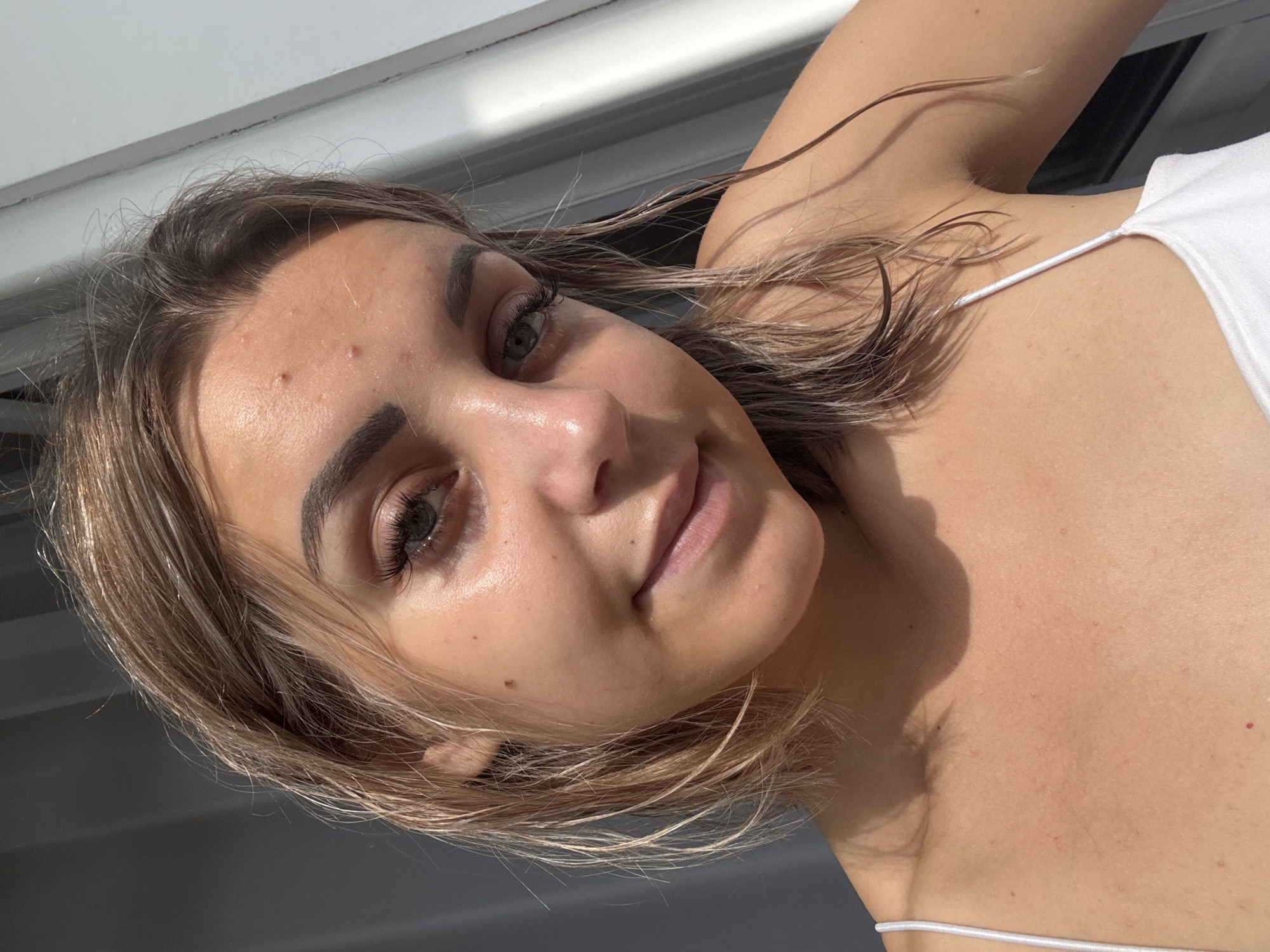 This is a close-up selfie of a young woman with light brown hair, styled in a bob cut that is slightly damp. She has fair skin with some minor blemishes and redness on her forehead and chest. Her eyes are visible, and she appears to be looking towards the camera with a slight smile. She is wearing a white, spaghetti-strap top. The lighting suggests natural sunlight, casting shadows on her face and chest. The background is out of focus, showing a plain, dark surface on the left and a lighter, possibly window or wall, surface on the right. The image is cropped to focus on her face, shoulders, and upper chest. There is no indication of the city of Kholmsk, Russia, or any specific urban environment in this image.