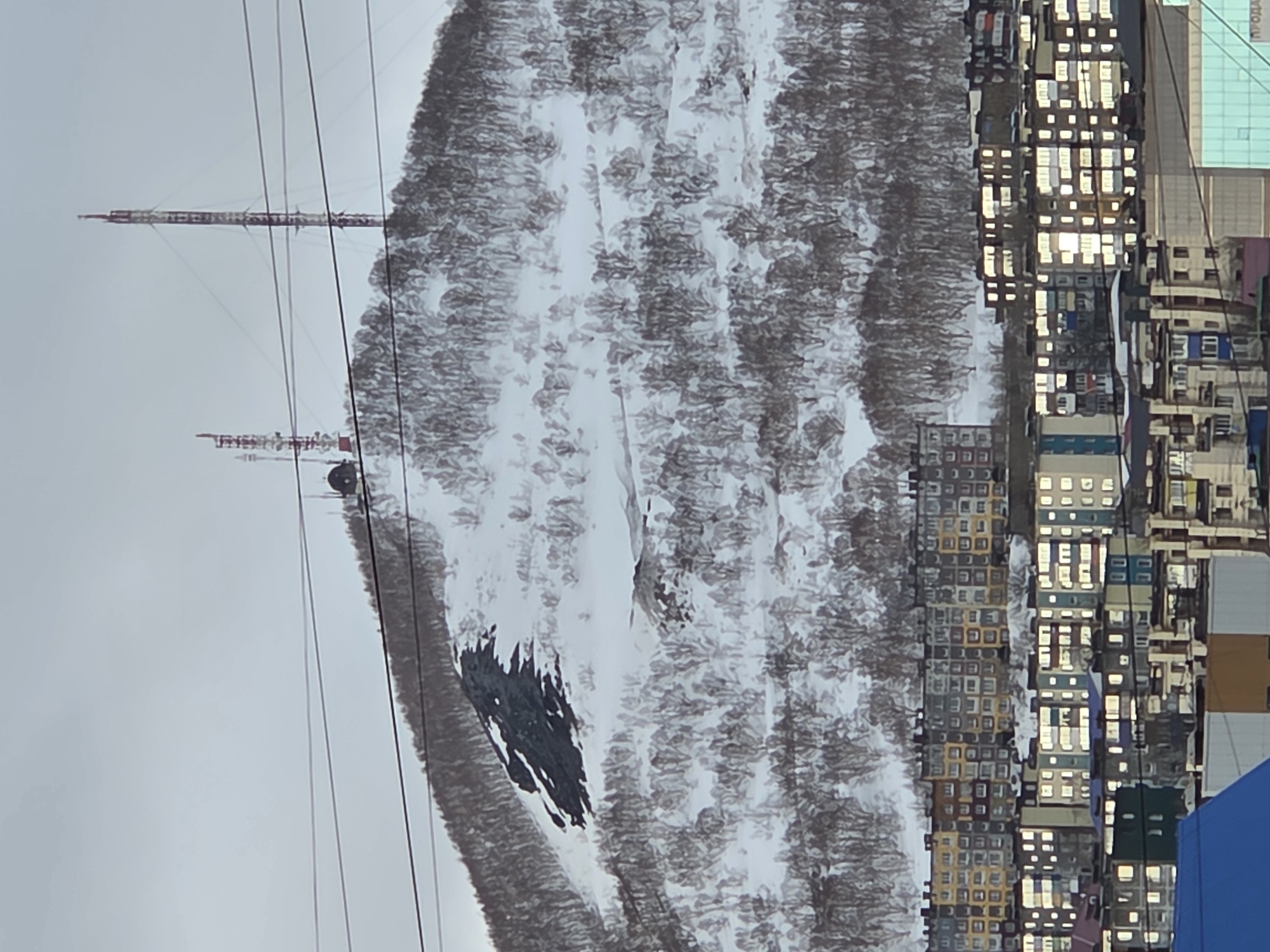 The image shows a winter scene. A snow-covered hill is visible, with bare trees interspersed. On the left, there are two towers with power lines stretching across the sky. Adjacent to the hill, on the right side of the image, is a dense cluster of buildings with many illuminated windows. The overall scene seems to depict an urban area nestled near a snow-covered hill.