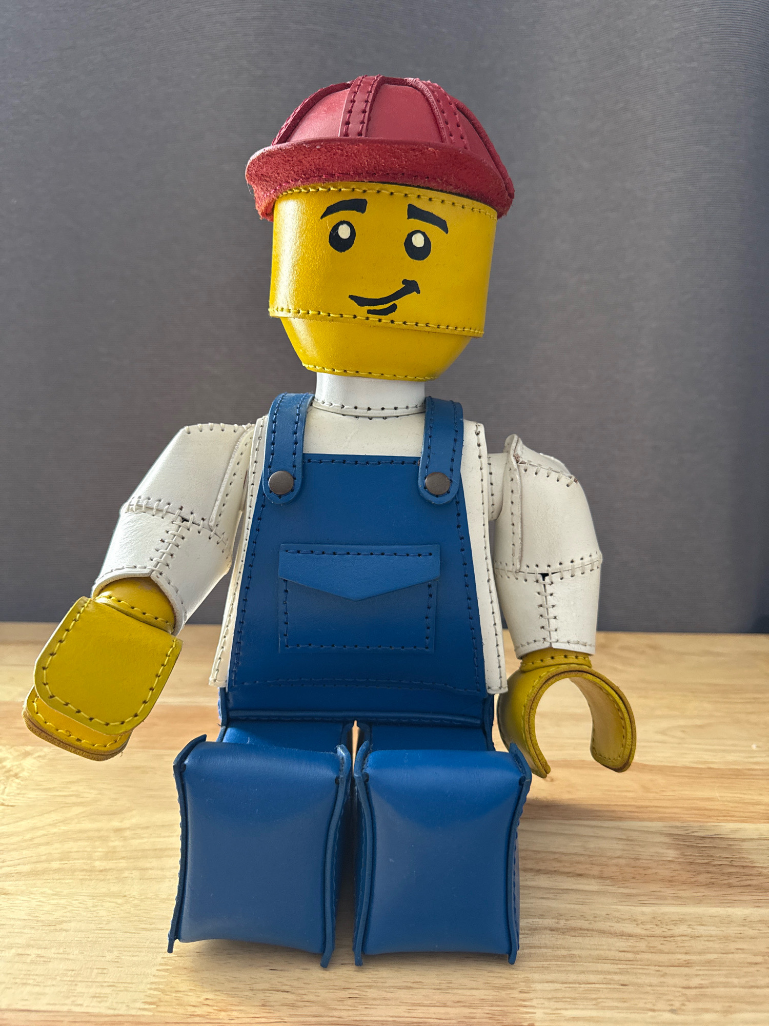An anthropomorphic leather figure resembling a LEGO minifigure stands upright on a light-colored wooden surface against a grey textured background. The figure has a yellow head with black eyes and a smiling mouth, and wears a red cap. Its body is covered by a white long-sleeved shirt and blue overalls with a bib pocket. The figure's arms and hands are yellow, and its legs are blue. The figure is positioned centrally in the frame, with its arms slightly extended. The lighting appears to be from above, casting minimal shadows. There is no indication of the location being New York City or any specific urban environment.