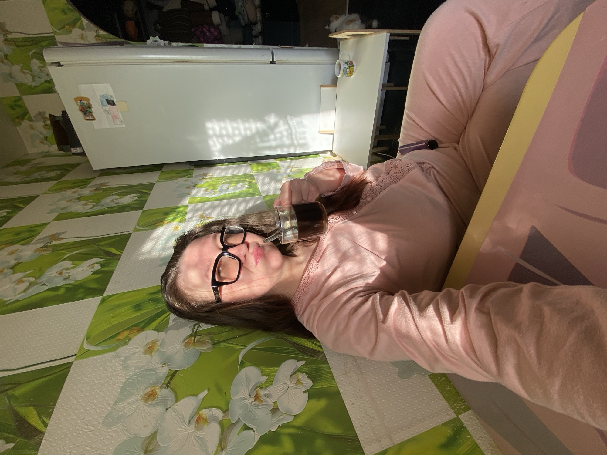 An adult female is depicted indoors, seated and angled towards the camera, holding a clear glass mug containing a dark beverage in her right hand. She wears black-framed glasses and pink loungewear with lace detail, her expression featuring a slight pout. The room's walls are covered with wallpaper displaying white orchids on a green and white checkered pattern, interspersed with textured white squares. To the right, a white refrigerator with attached magnets is visible, positioned next to a small white table with two chairs, one of which holds a patterned mug. A white and orange cat is partially visible on the dark floor near the table. Strong sunlight enters from the left, casting distinct striped shadows on the refrigerator and adjacent wall. This scene is located within a residence in the city of Chaikovsky, Russia.