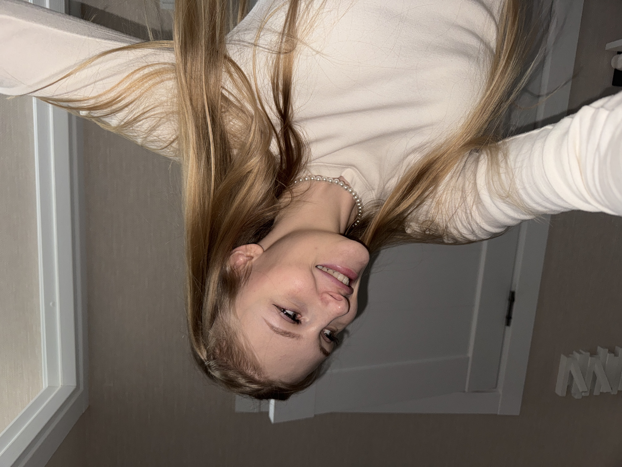 A young woman with long blonde hair is pictured from the chest up, taken from a low angle. She is wearing a pearl necklace and a cream-colored long-sleeved shirt. The background shows a white door frame and a light beige wall. The lighting is dim, suggesting an indoor setting, possibly a home or apartment. There is no visible indication of the specific location in Perm, Russia.